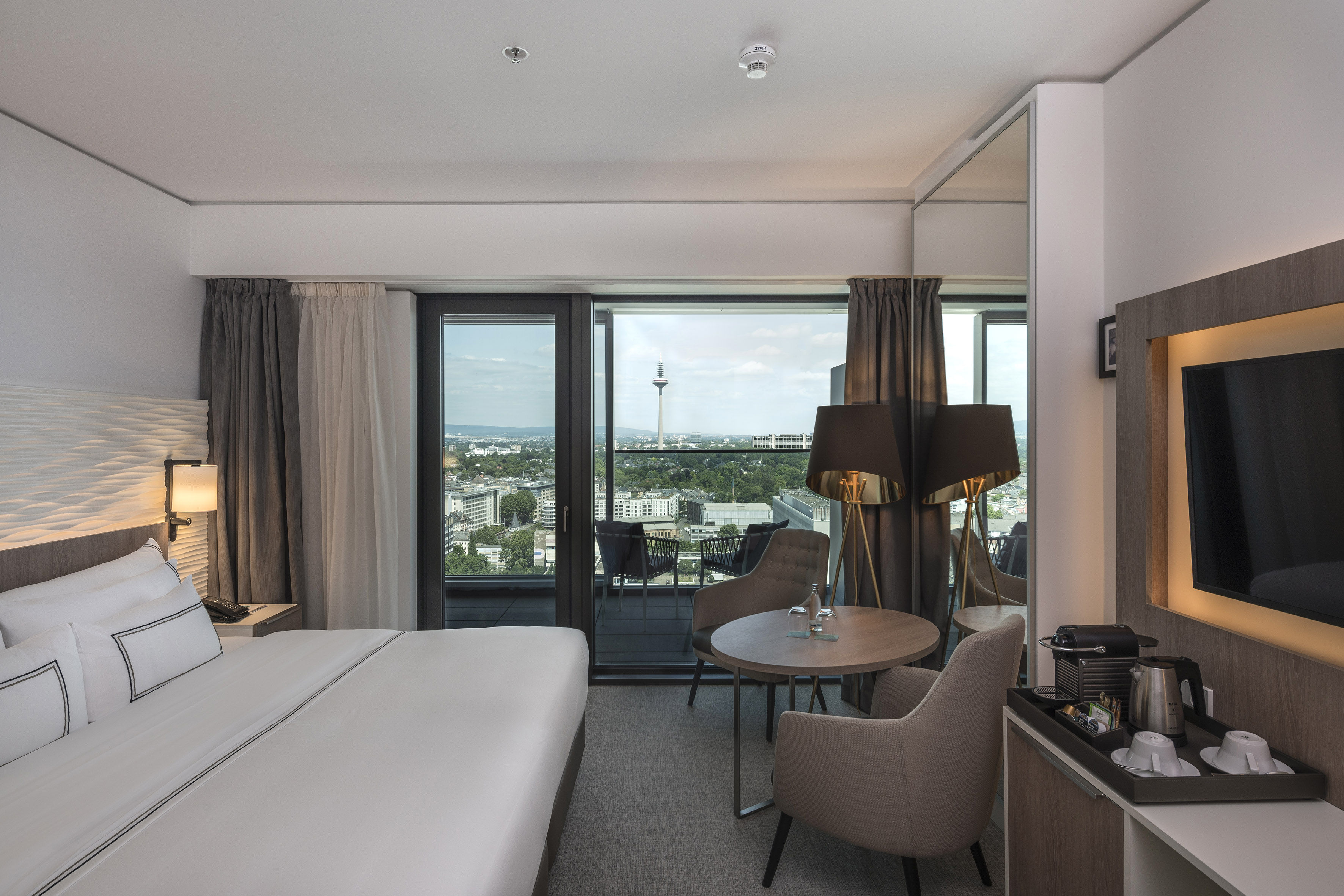 Spacious hotel room with bed, city view balcony, and Düsseldorf's Rheinturm.