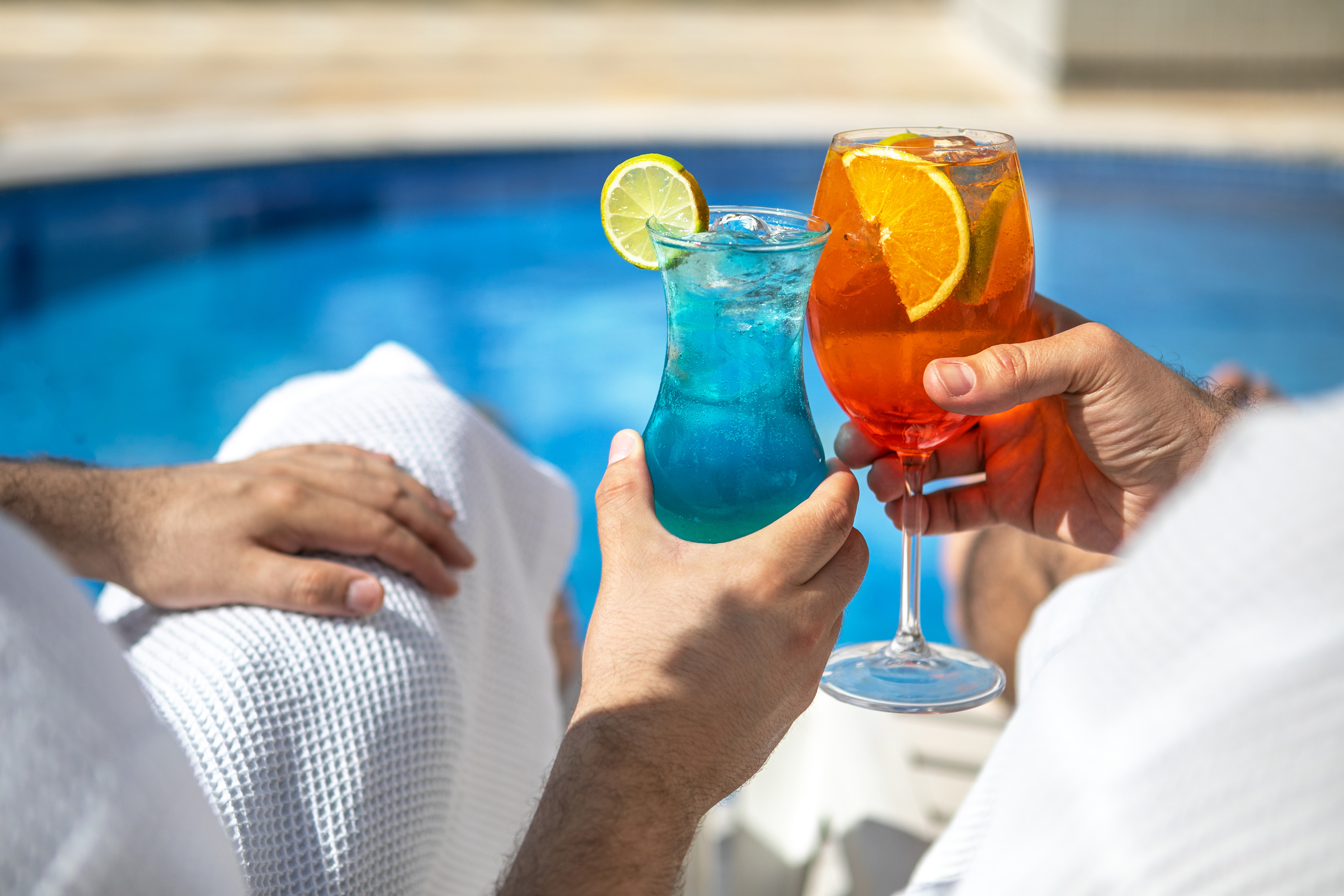 Hands holding blue and orange tropical cocktails by a swimming pool.