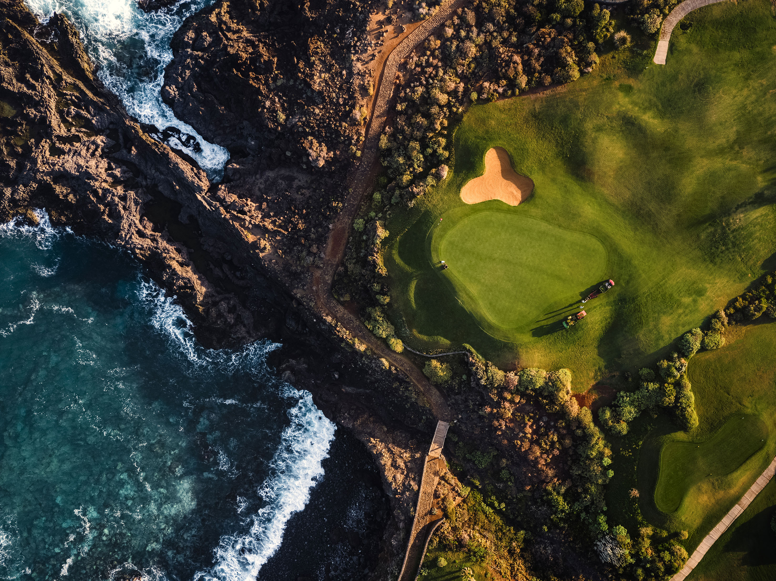 Oceanfront golf course aerial with green, sand bunker, rocky shore, and waves.