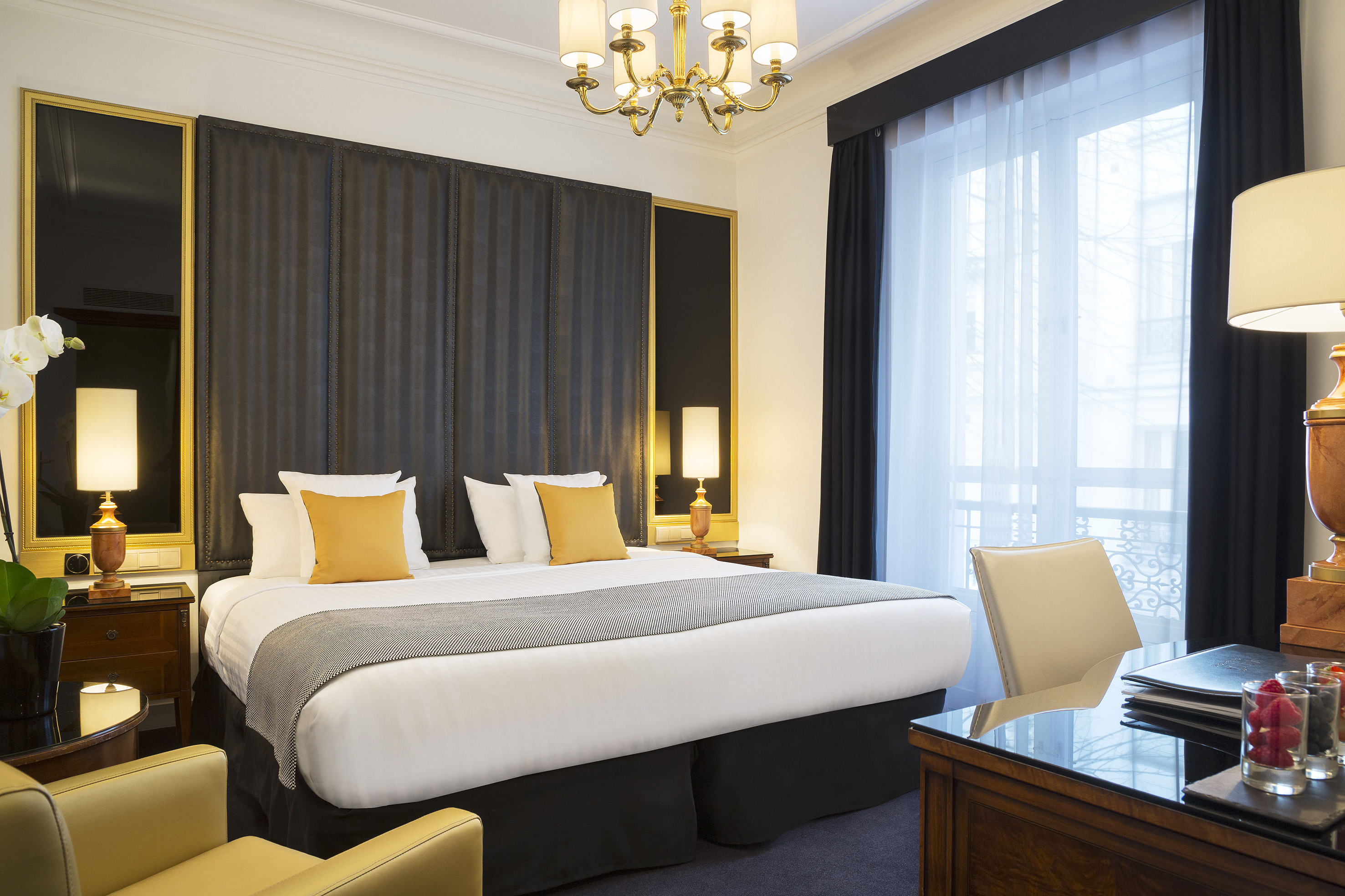Elegant hotel suite with king bed, chandelier, desk, and large window.