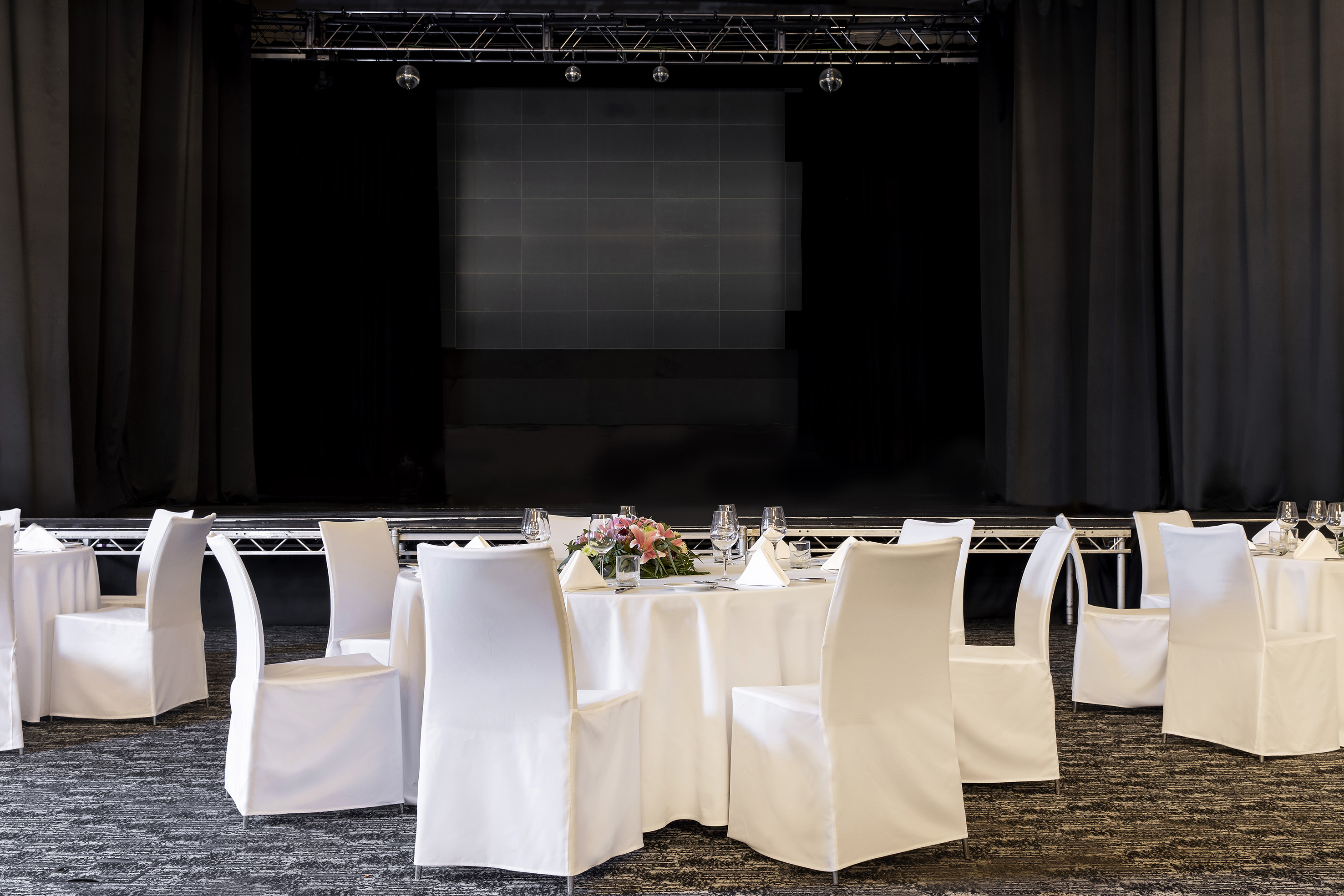 Banquet hall with round tables, white chair covers, and a stage backdrop.