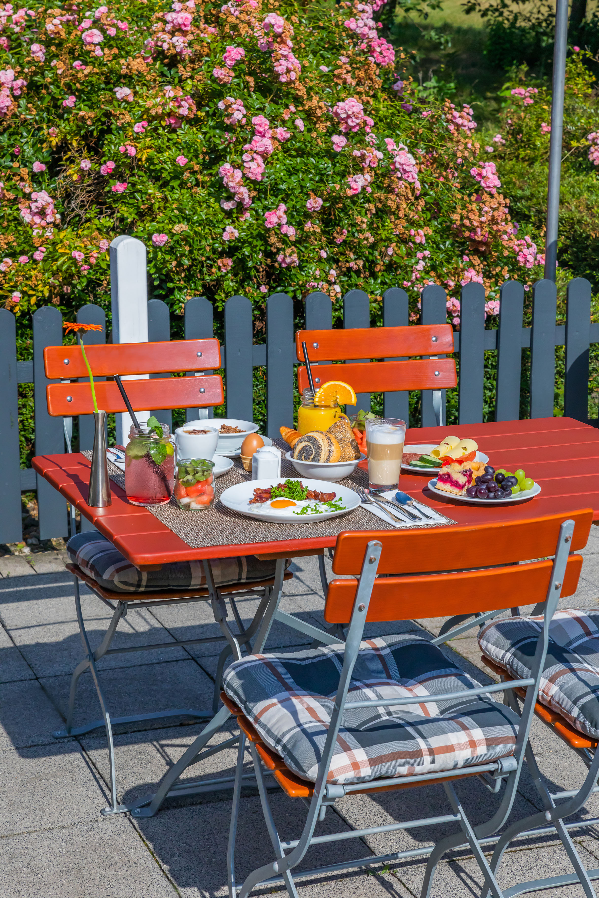 Sunny outdoor breakfast on red table with eggs, pastries, juice, and pink roses.