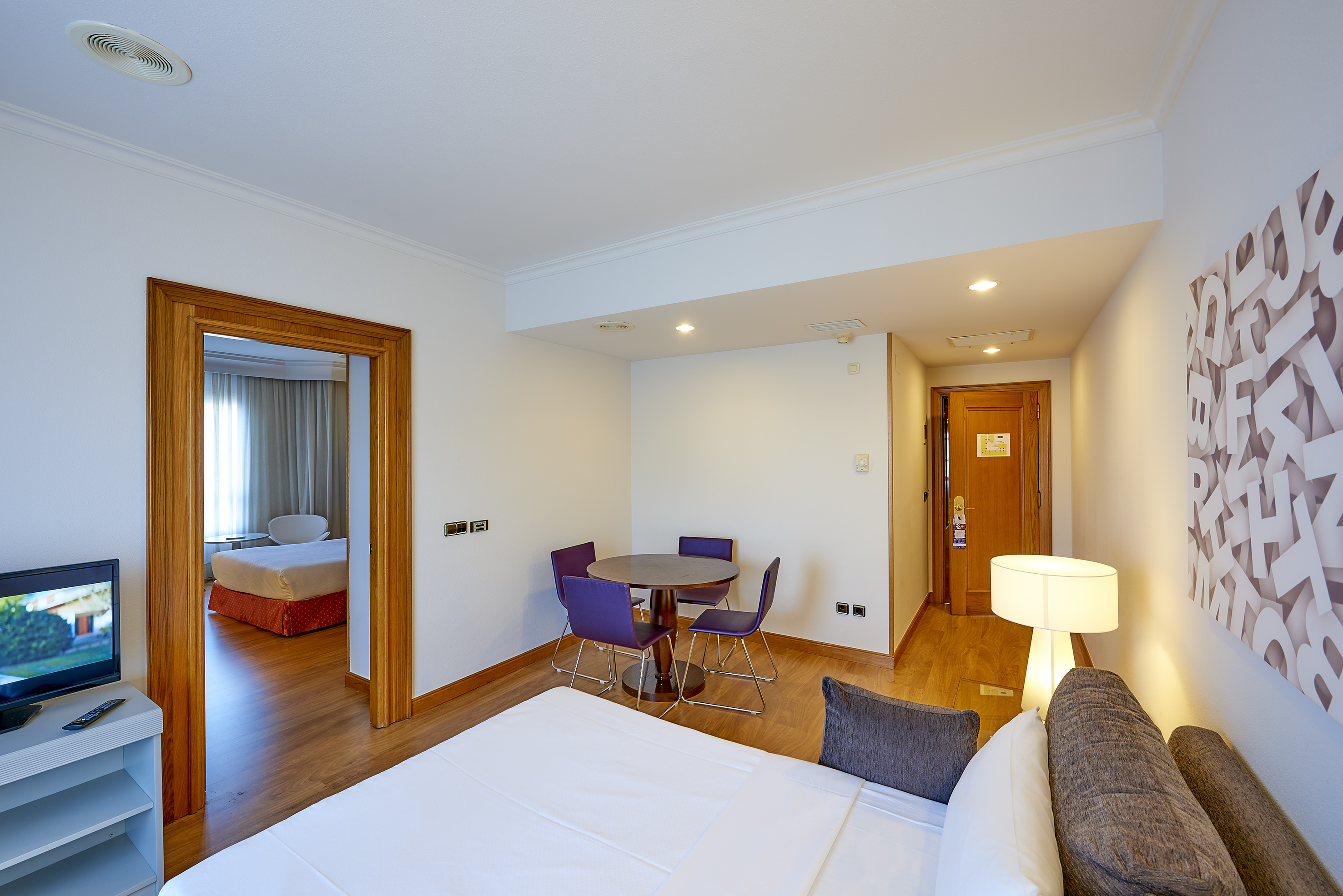 Hotel suite with bed, dining table with purple chairs, and connected bedroom.