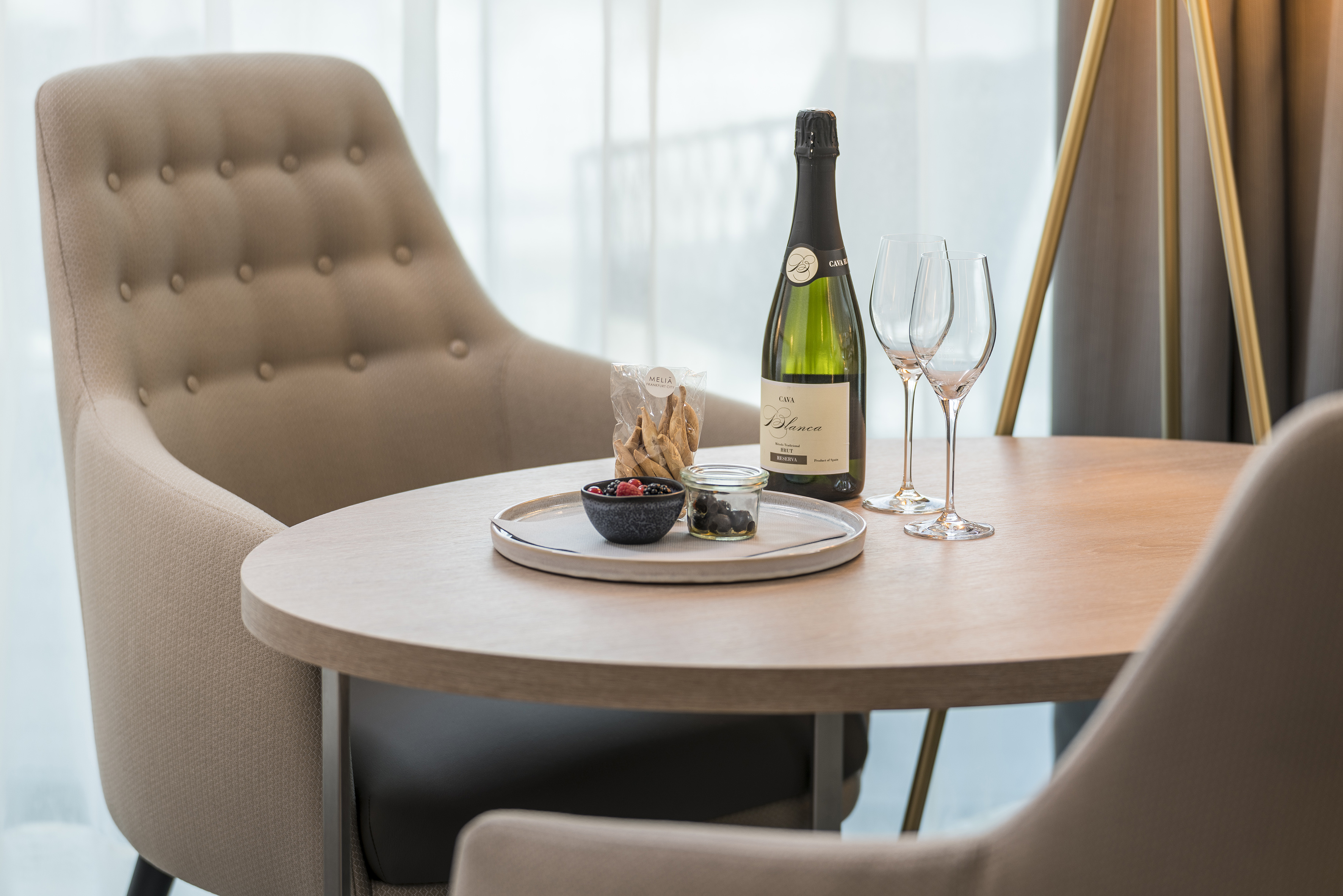 Hotel room table with Cava, wine glasses, berries, and snacks between beige chairs.