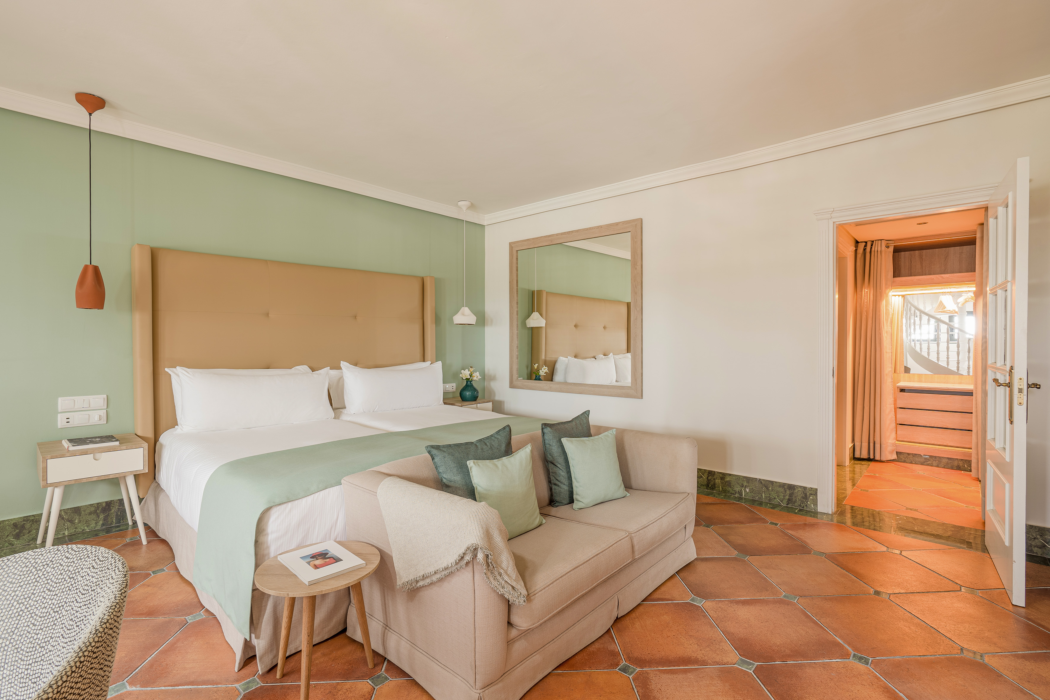 Hotel bedroom with two beds, beige sofa, terracotta tiles, and green accent wall.
