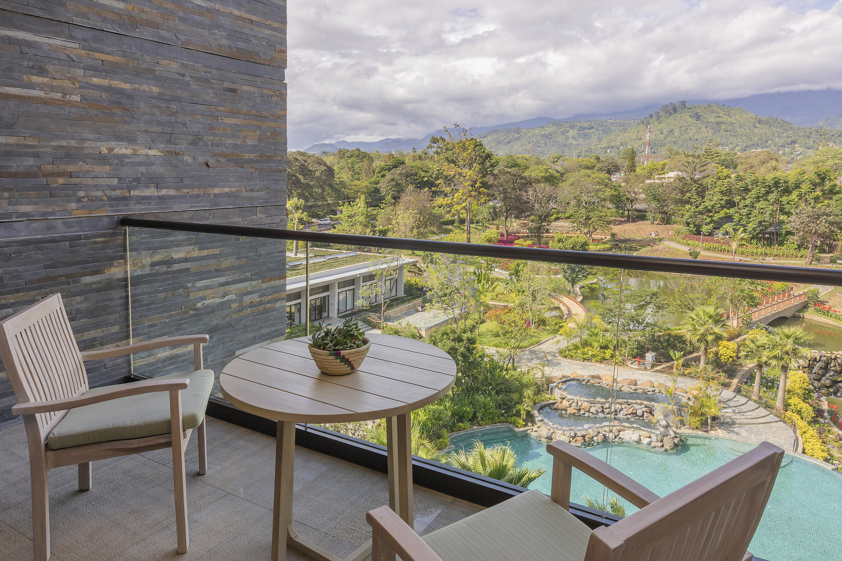 Hotel balcony with outdoor seating overlooking tropical pool, gardens, and mountains.