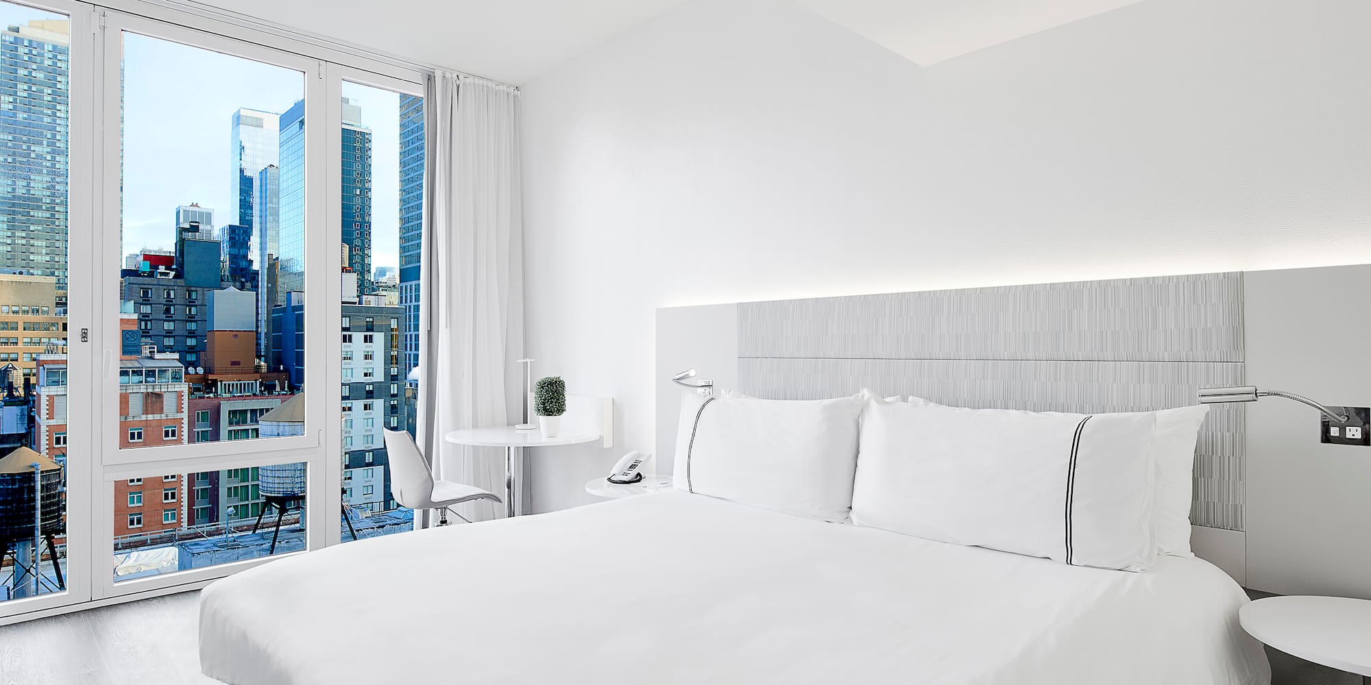 a white bed with white sheets and a table and chairs in a room with a city view