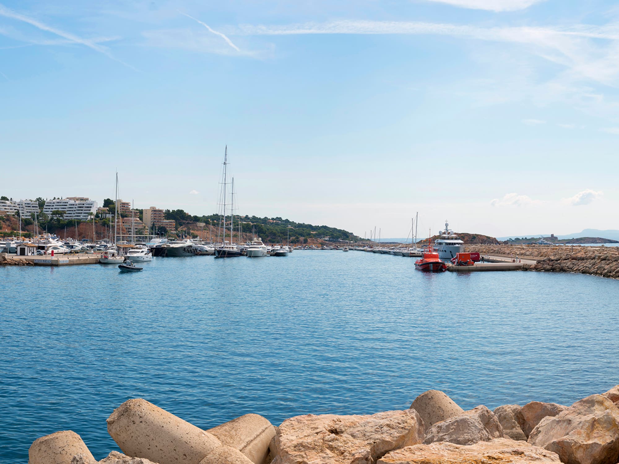 Puerto deportivo mediterráneo con yates, rompeolas de roca y costa urbana.