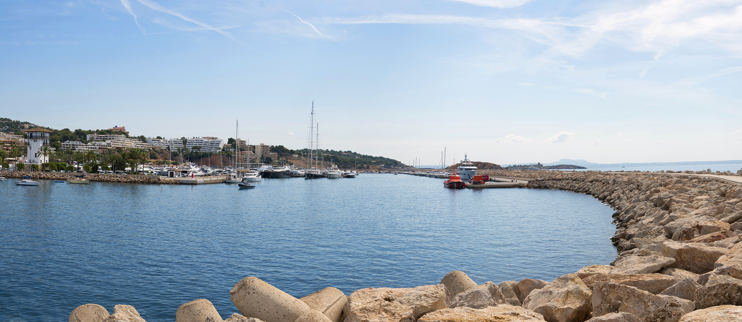 Puerto deportivo mediterráneo con yates, rompeolas de roca y costa urbana.