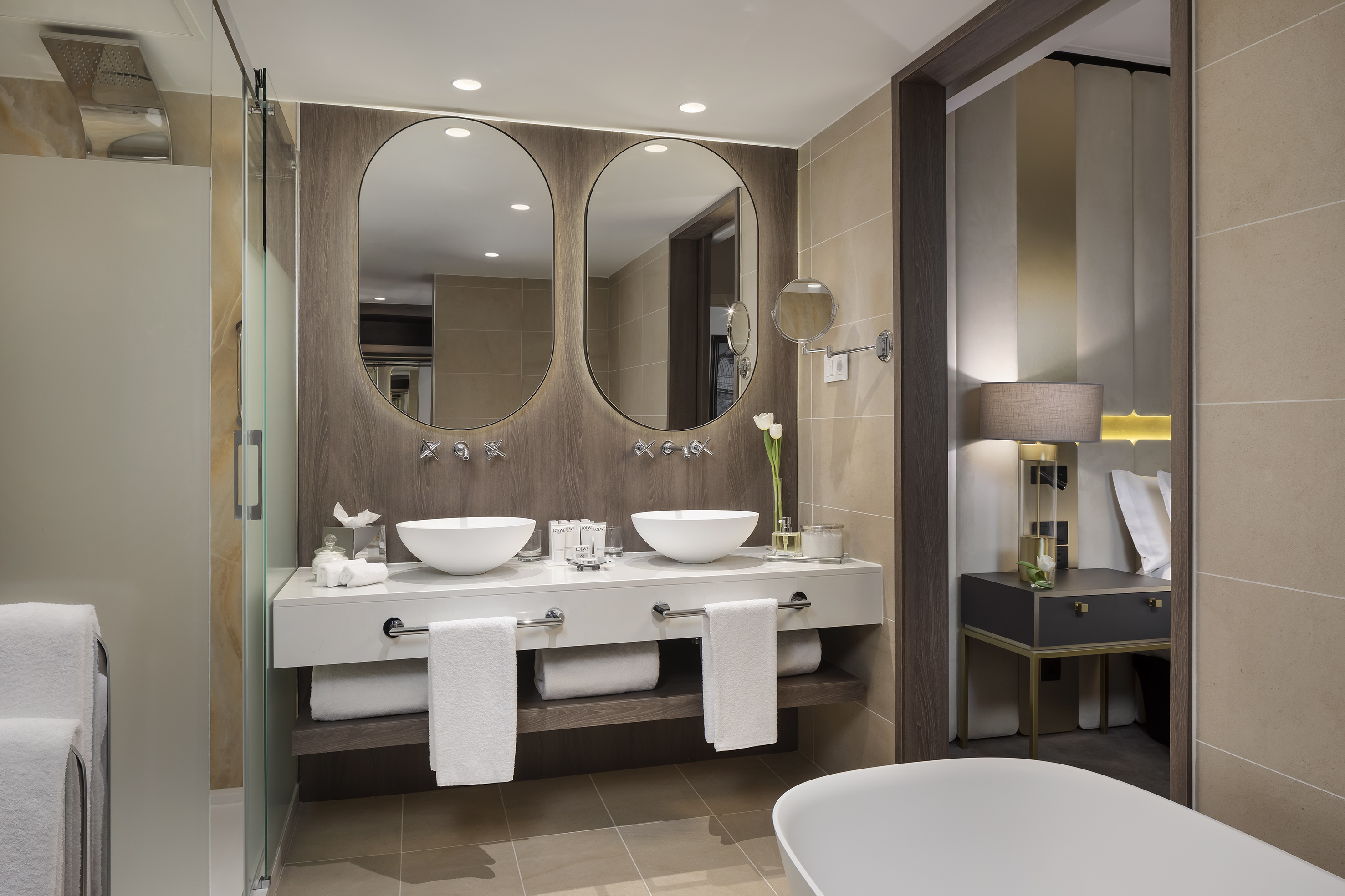 Modern hotel bathroom: dual sinks, oval mirrors, shower, and connected bedroom.
