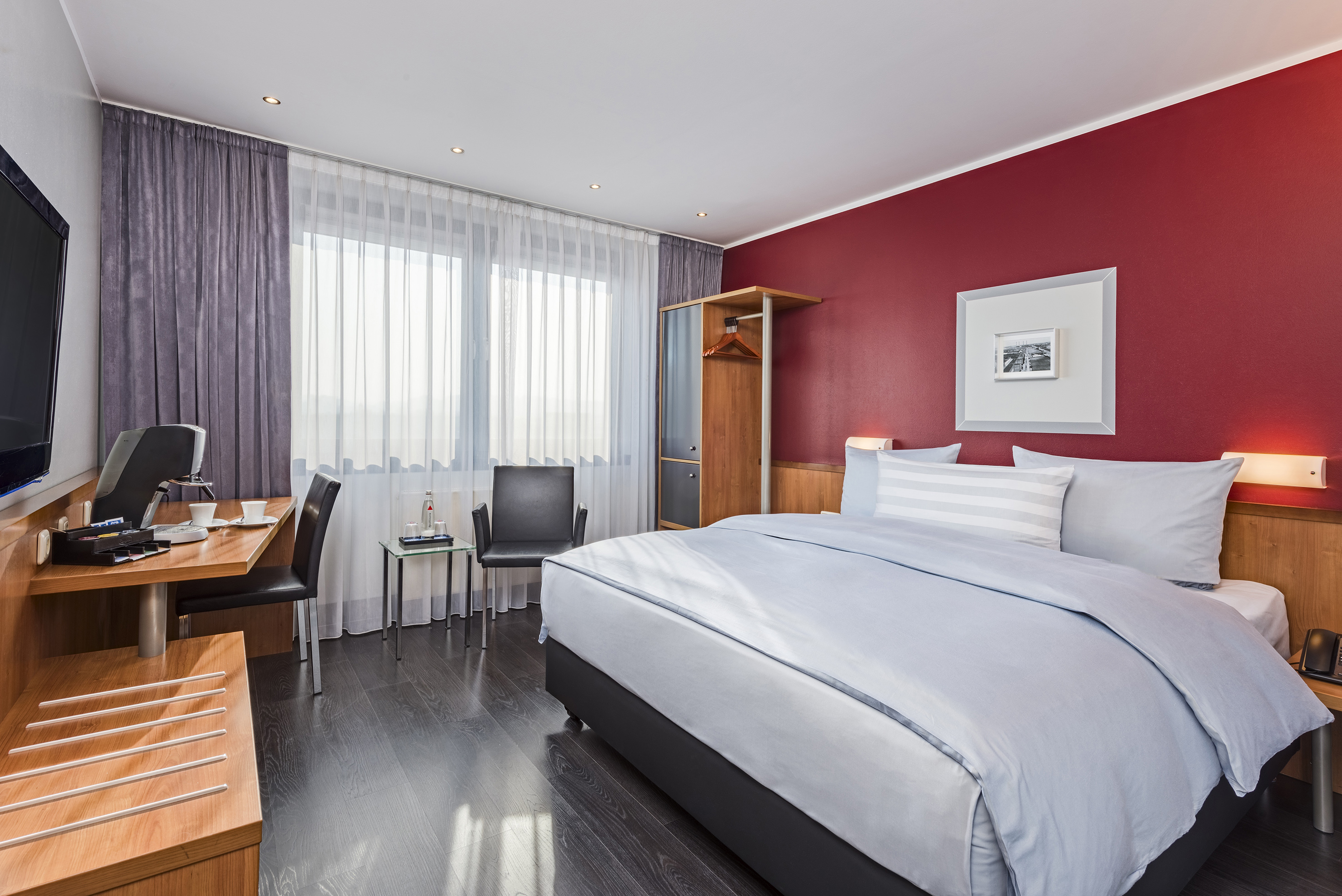 Modern hotel room with blue bedding, desk, red accent wall, and window.