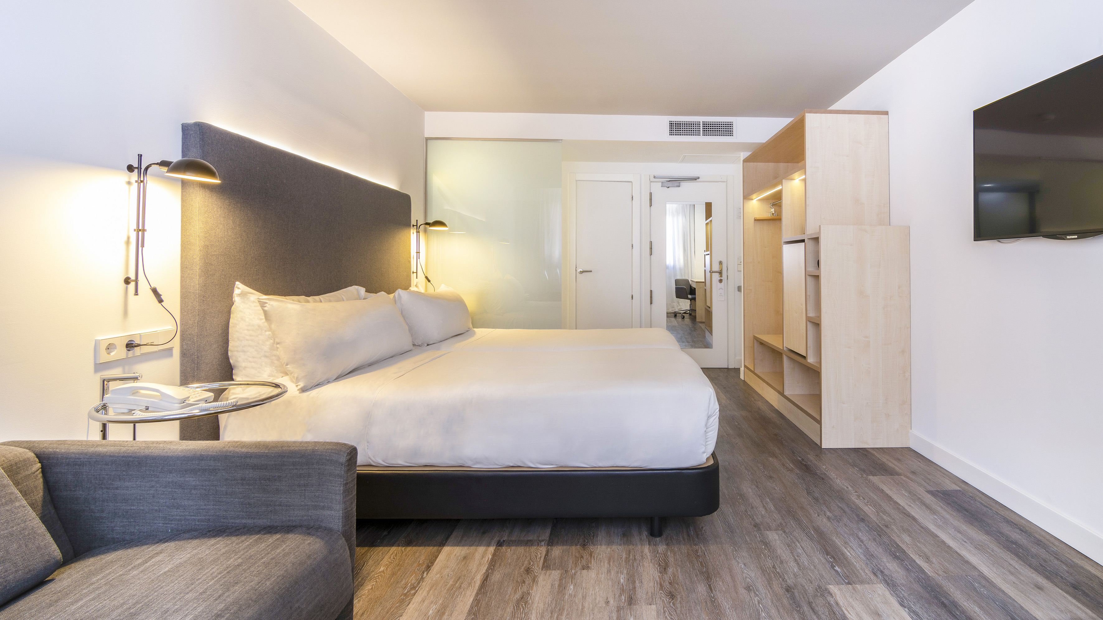 Modern hotel room with two beds, gray sofa, flat-screen TV, and wood floor.