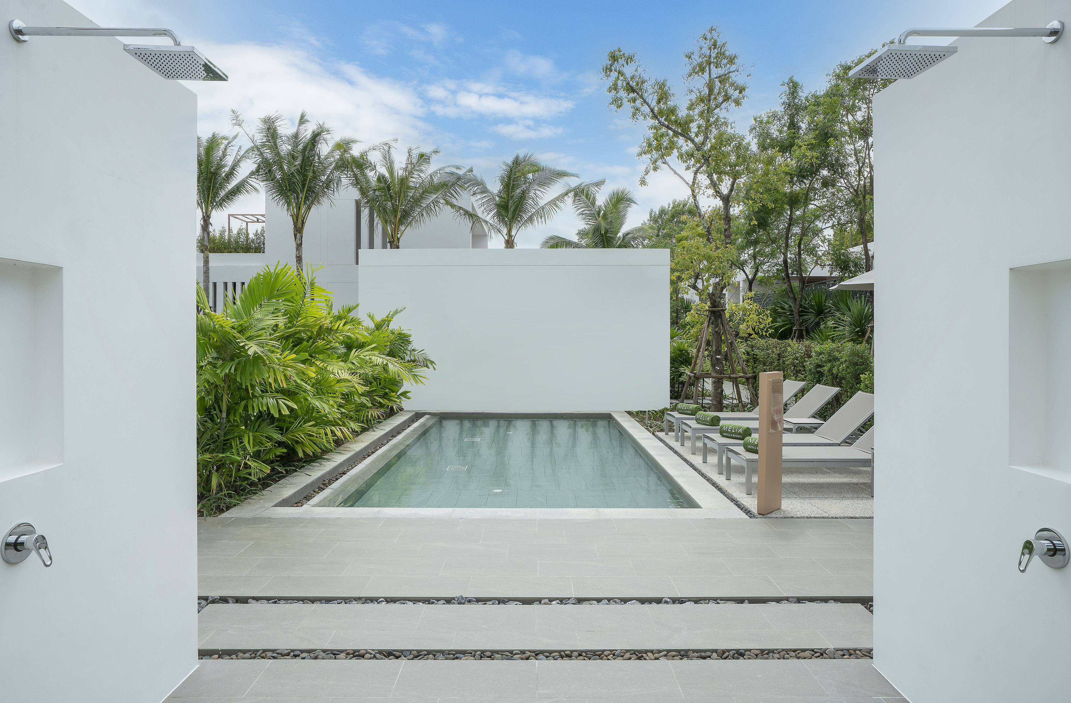 Tropical resort pool area with sun loungers, outdoor showers, and lush plants.