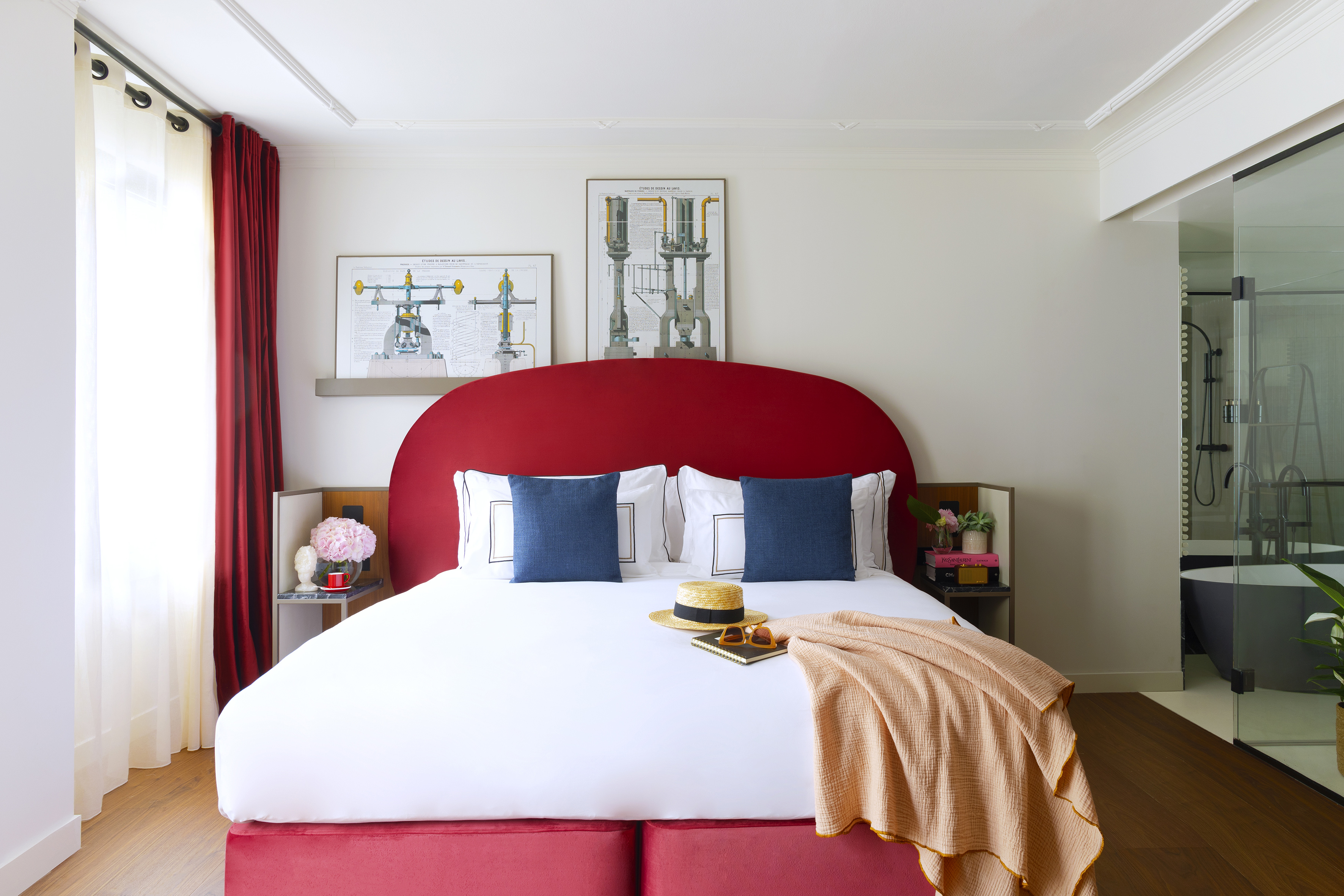 Modern hotel bedroom: red bed, white linens, blue pillows, glass bath partition.