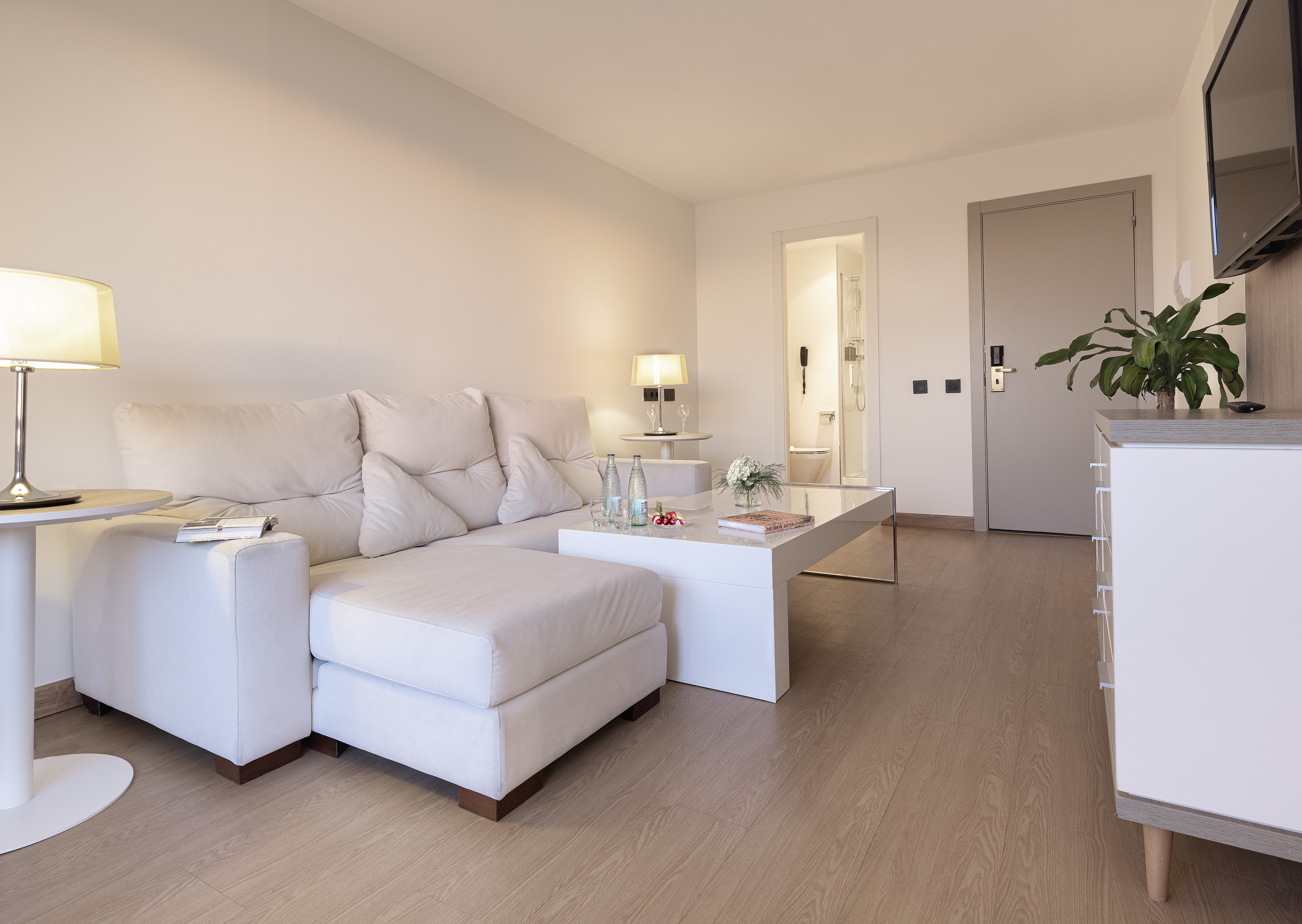 Modern hotel suite living room with white sectional sofa and wood floor.