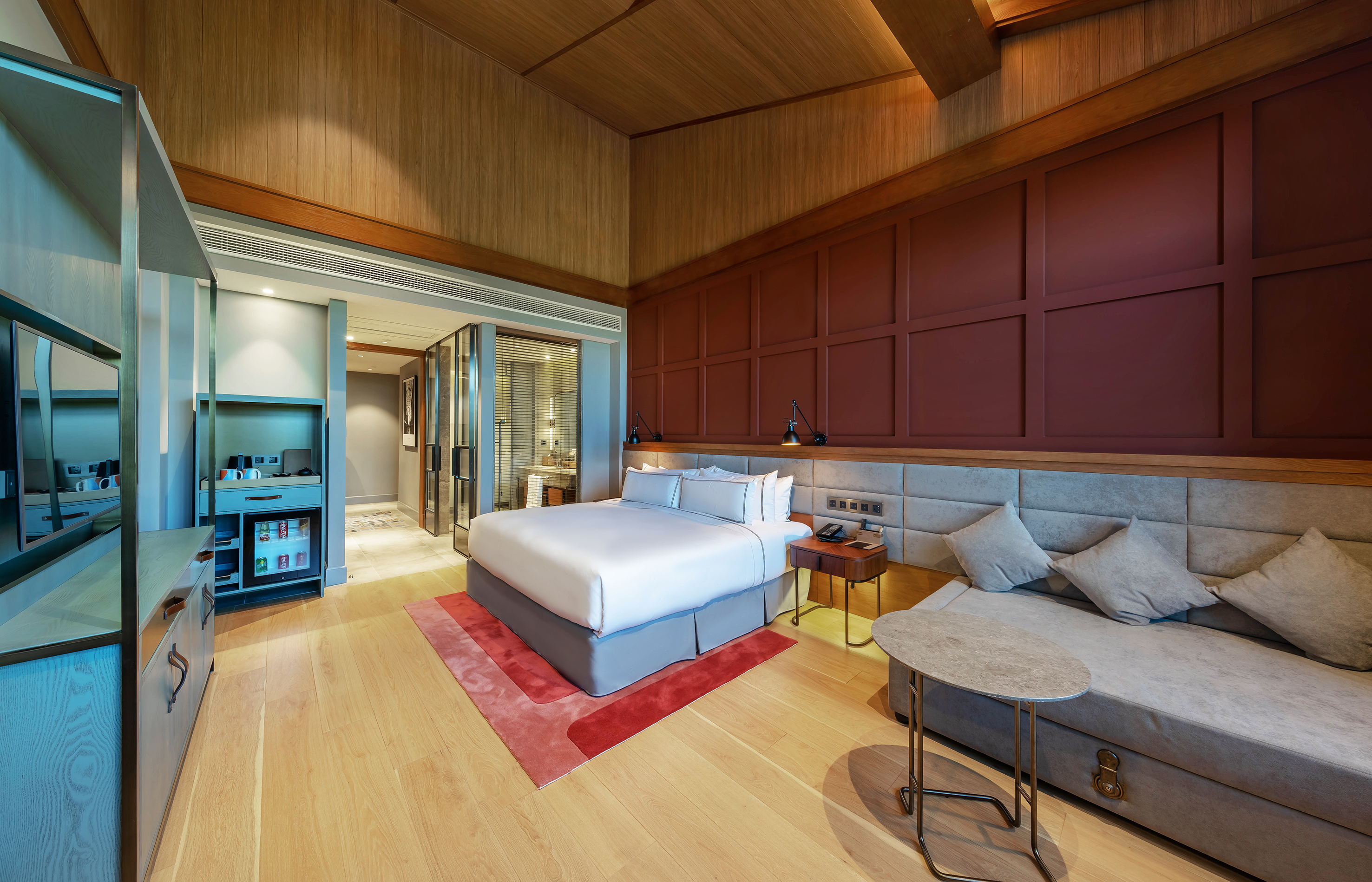 Modern luxury hotel room with large bed, red paneled wall, wooden ceiling, and sofa.
