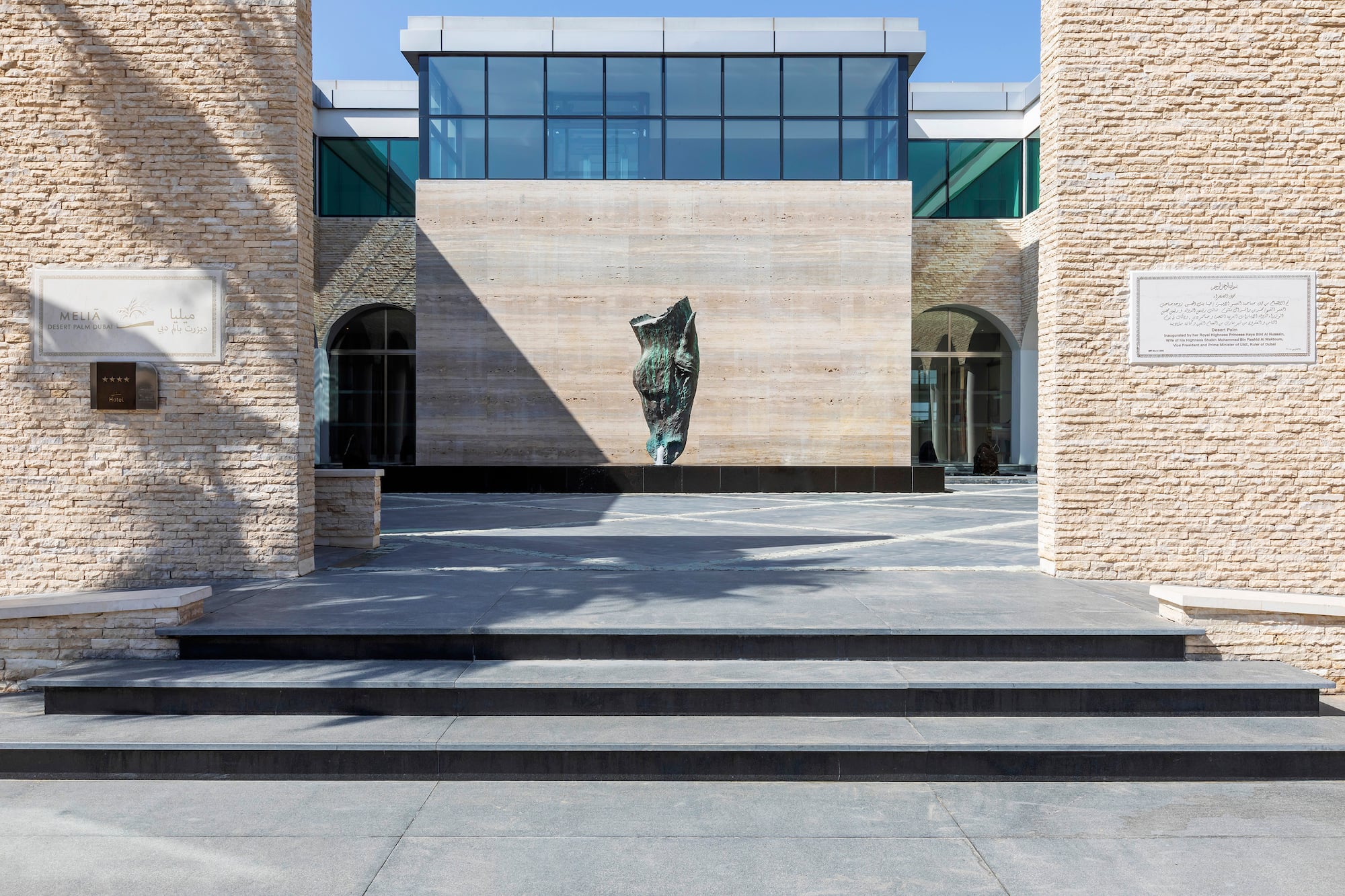 Meliá Desert Palm Dubai hotel facade: stone walls, contemporary sculpture, glass windows.