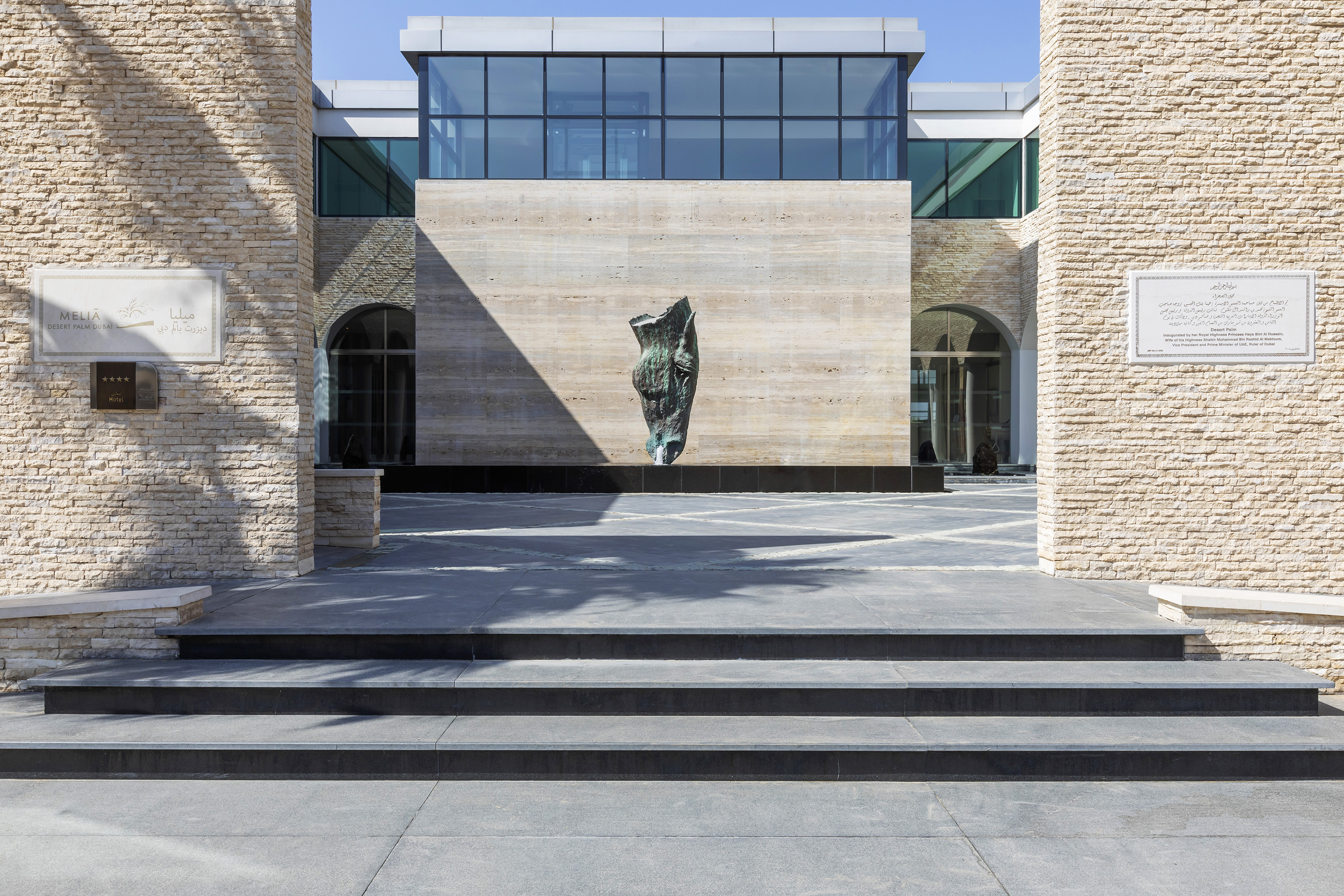 Meliá Desert Palm Dubai hotel facade: stone walls, contemporary sculpture, glass windows.