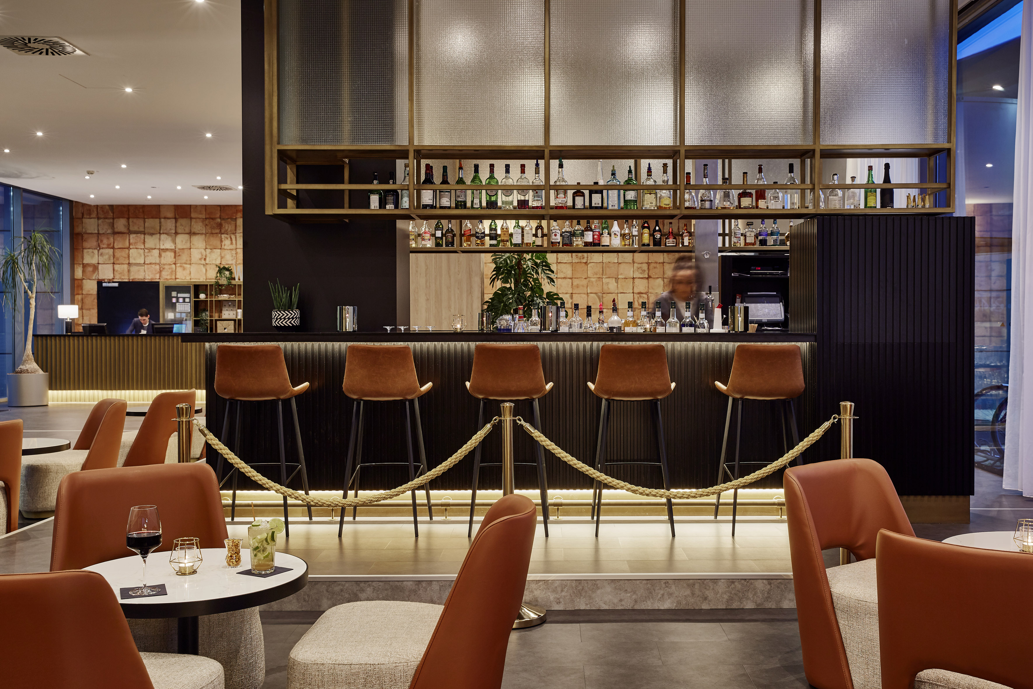 Modern hotel bar interior with liquor bottles, brown bar stools, and lounge chairs.