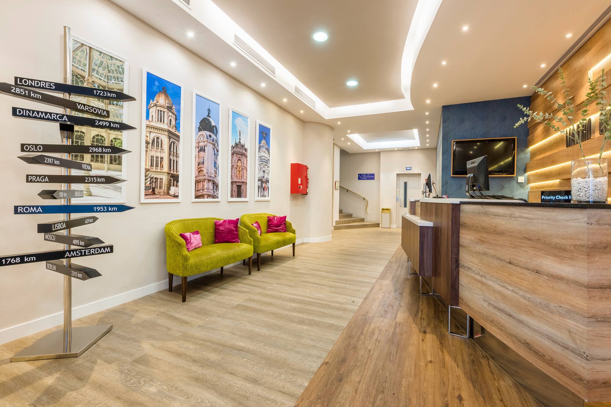 Modern hotel lobby with reception desk, green armchairs, and a directional signpost.