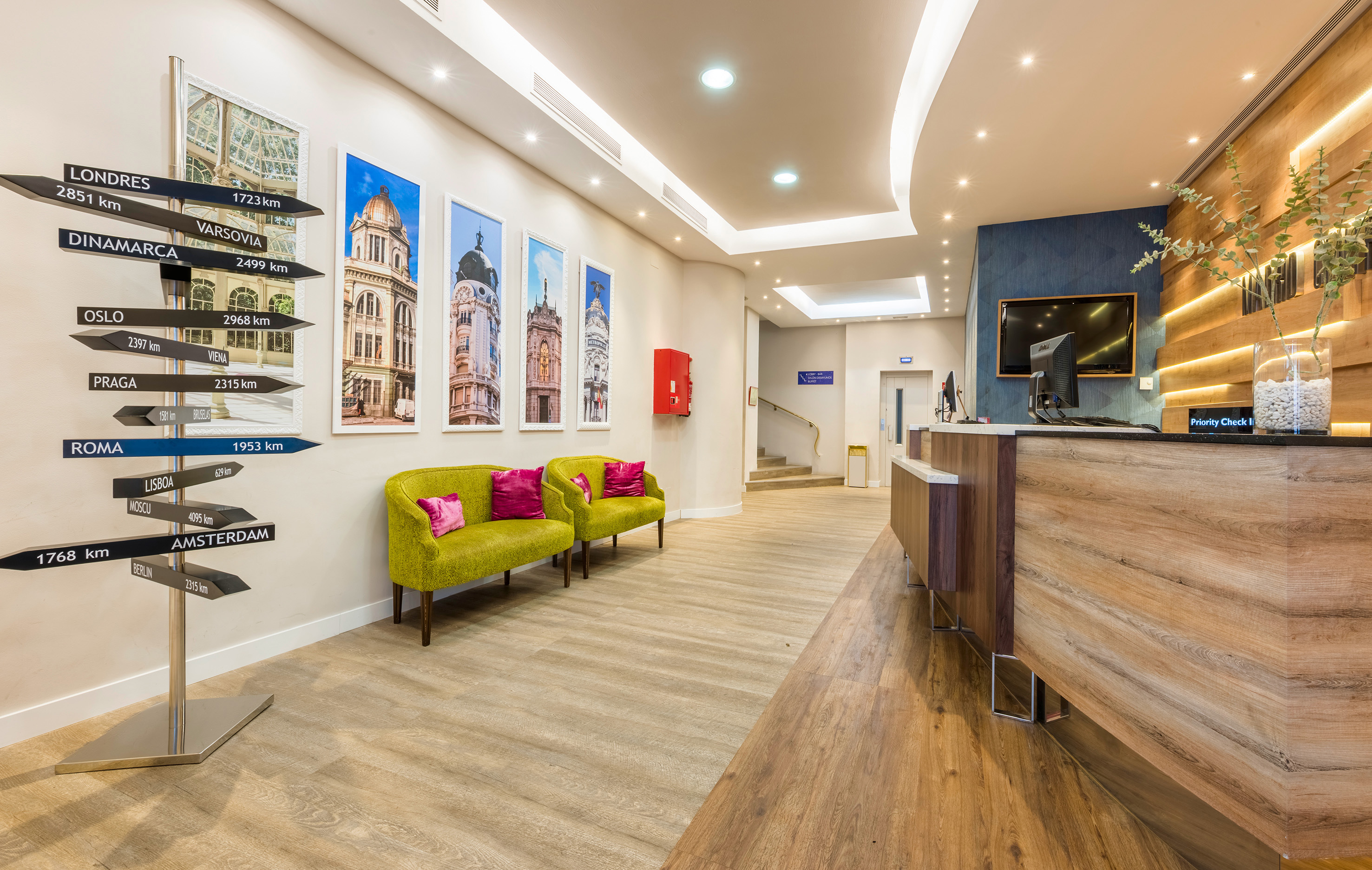 Modern hotel lobby with reception desk, green armchairs, and a directional signpost.