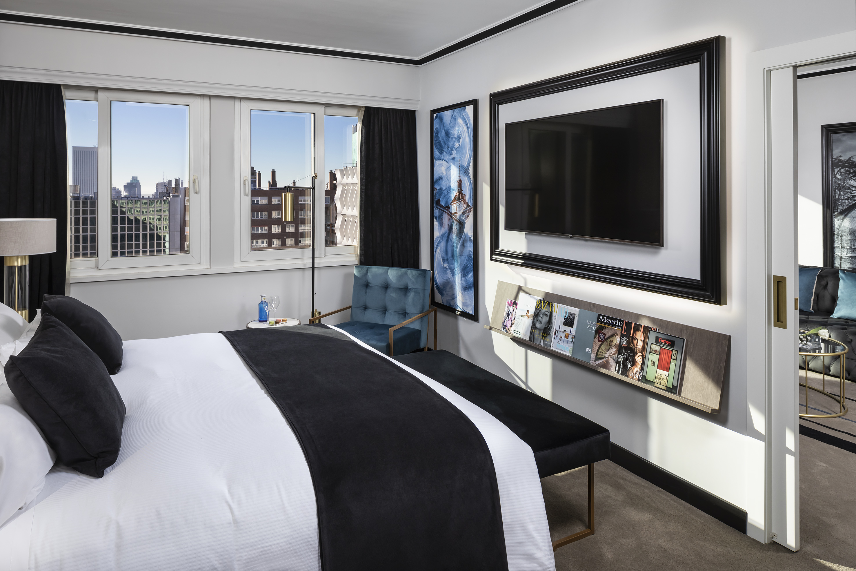 Stylish hotel bedroom with city skyline view, comfortable bed, TV, and blue armchair.
