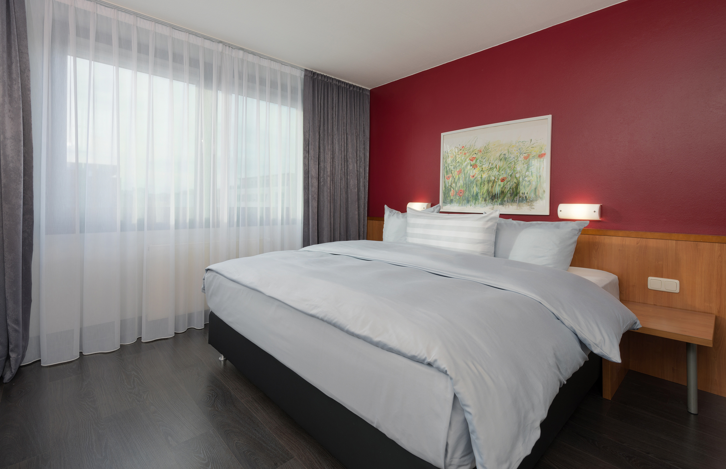 Modern hotel bedroom with king bed, grey bedding, red wall, and window.