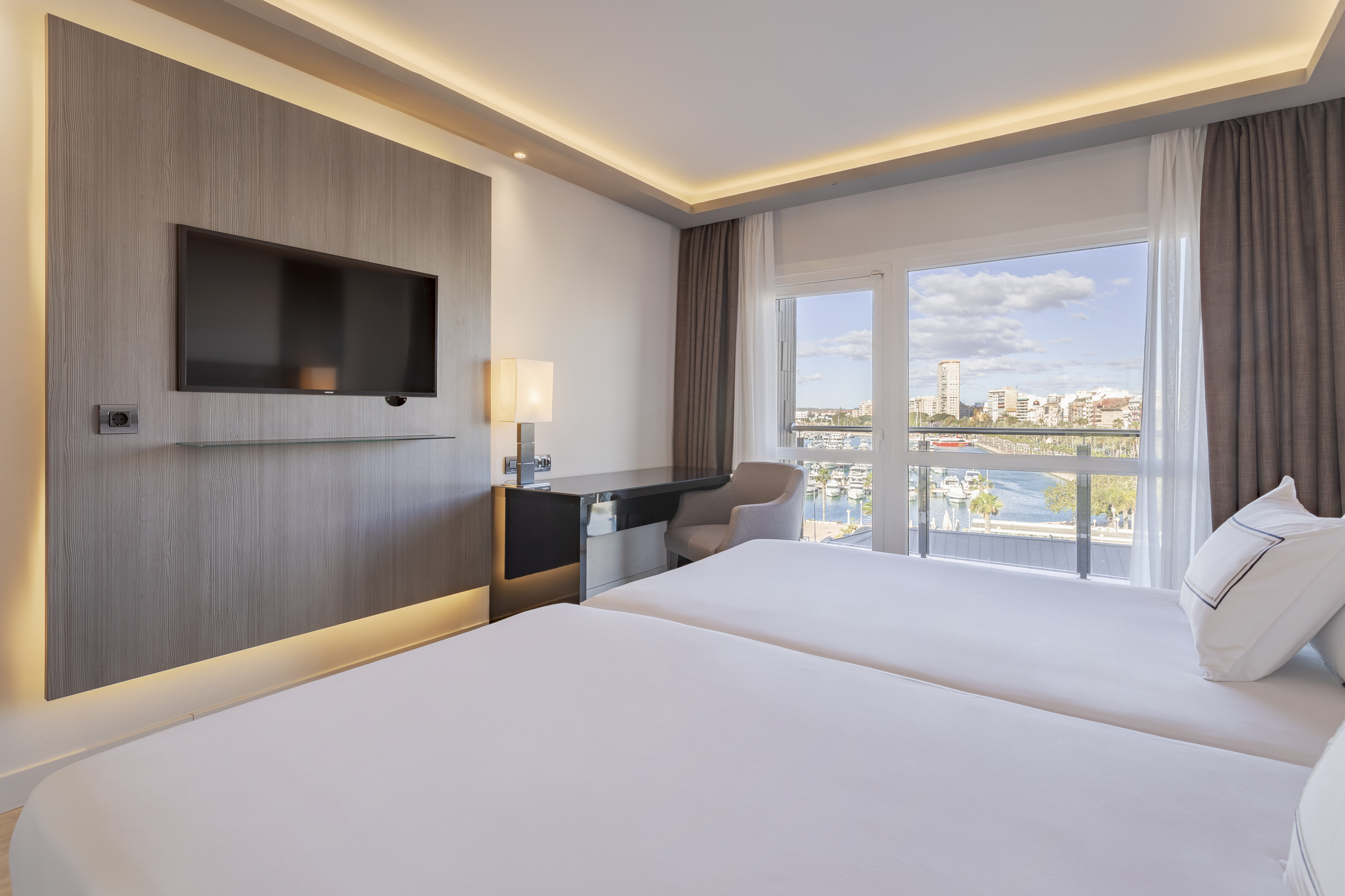 Modern hotel room with twin beds, TV, desk, and marina view.