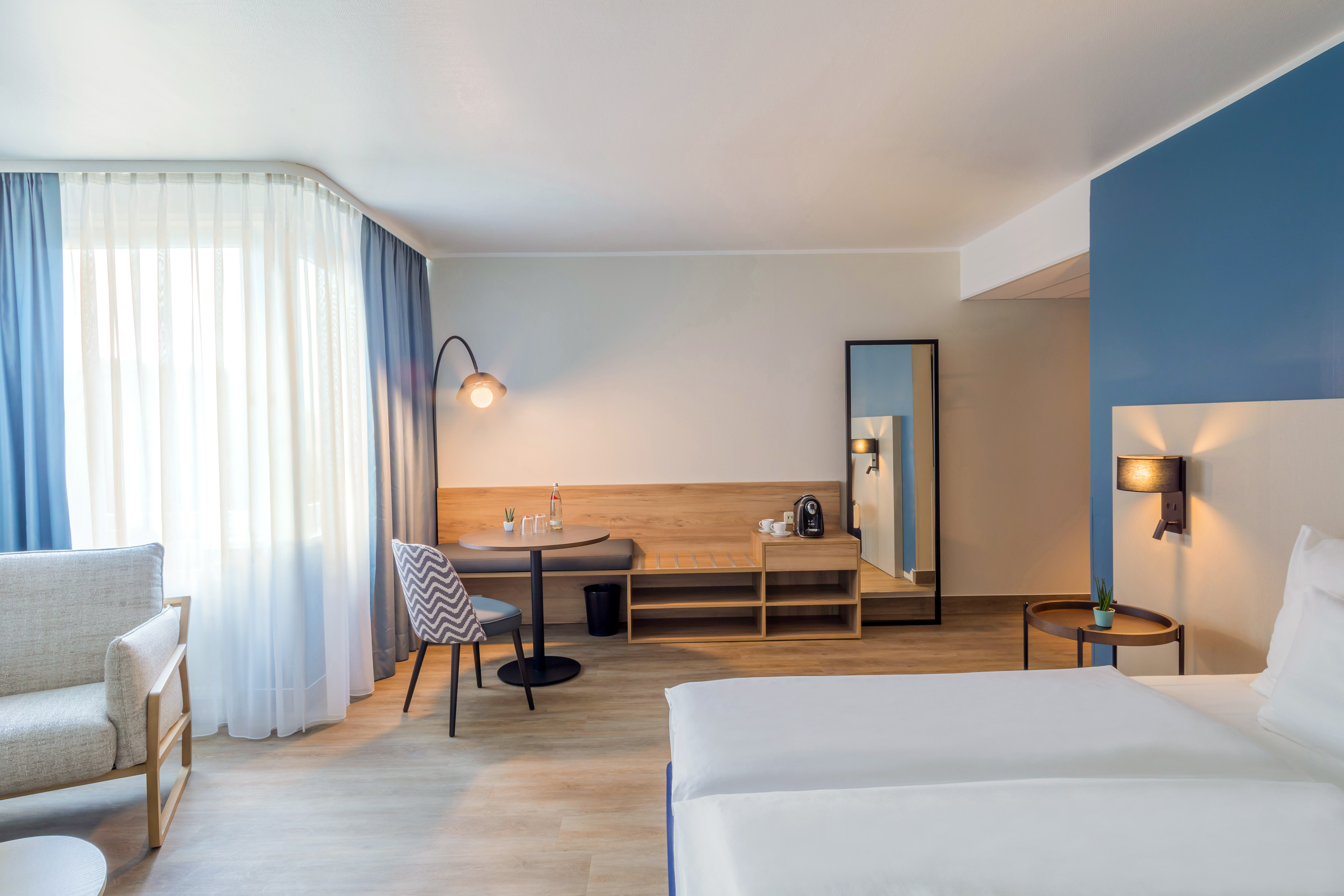 Modern hotel room with white bed, blue accent wall, and wooden dining area.