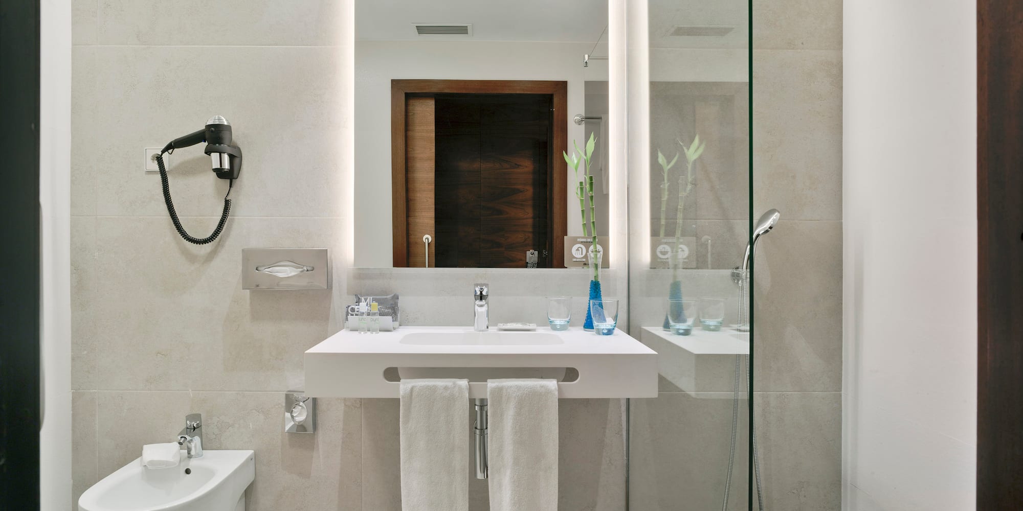 Elegante baño de hotel con lavabo, espejo iluminado, bidet y ducha acristalada.