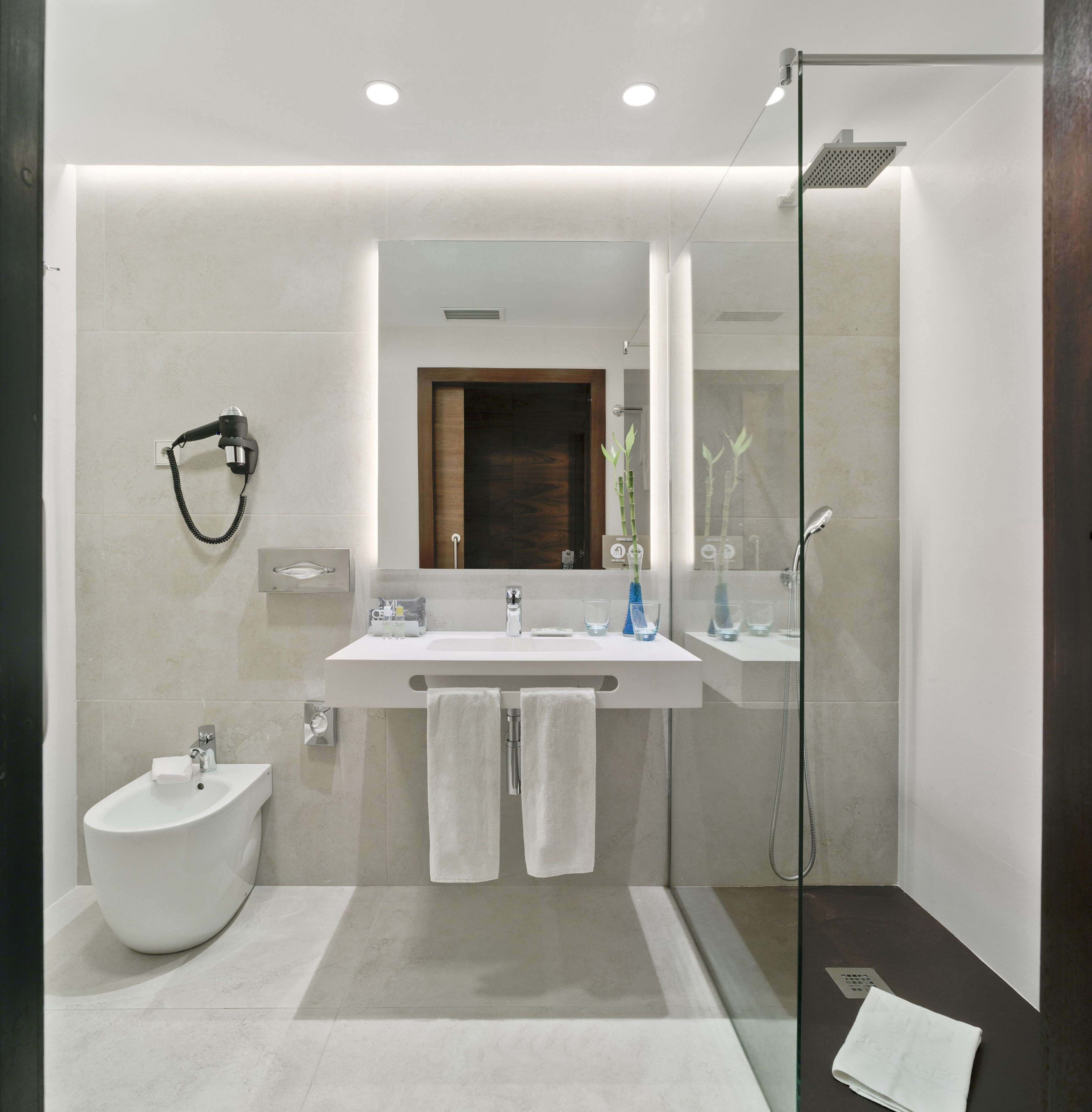 Elegante baño de hotel con lavabo, espejo iluminado, bidet y ducha acristalada.