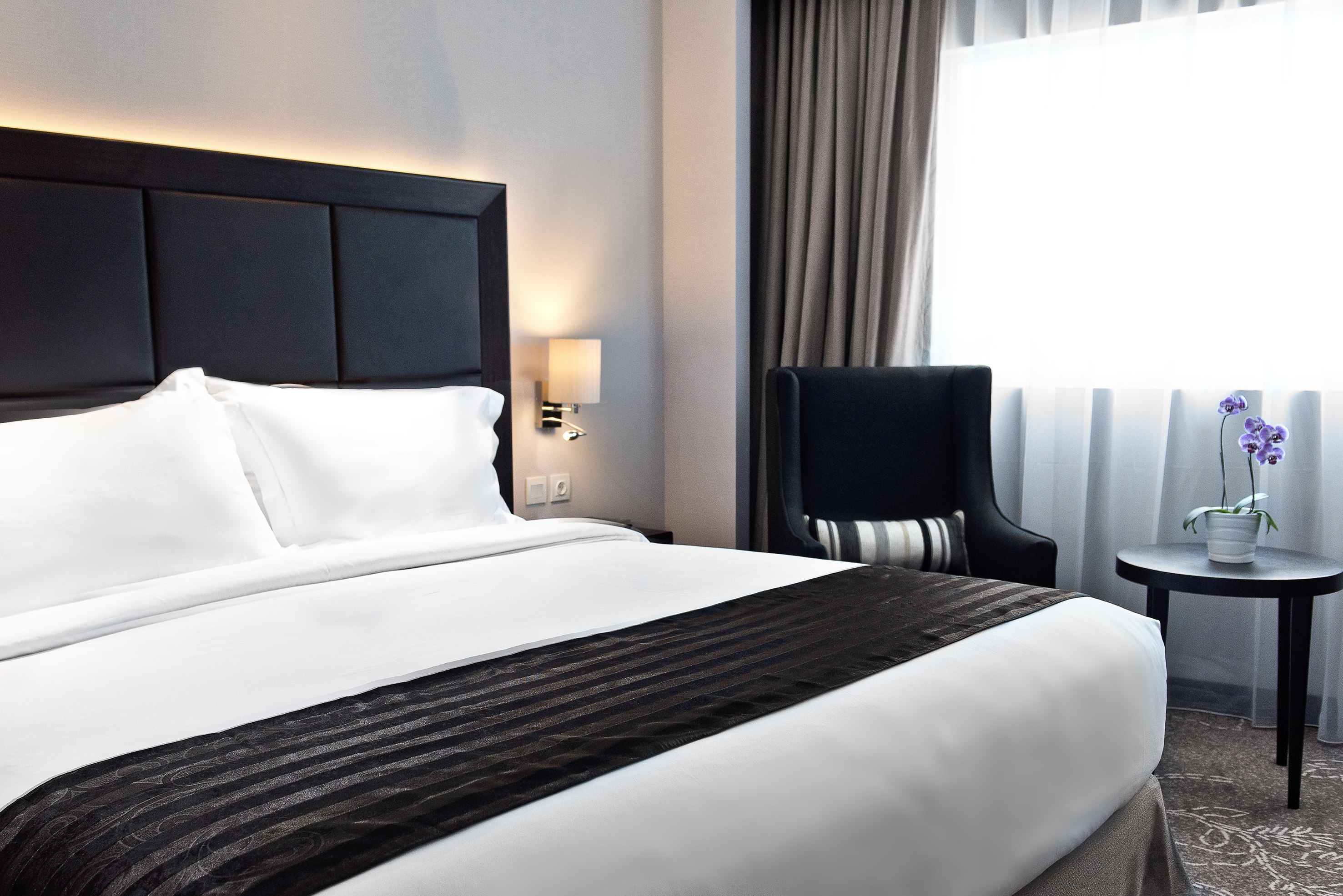 Modern hotel bedroom with white bed, black headboard, armchair, and orchid.