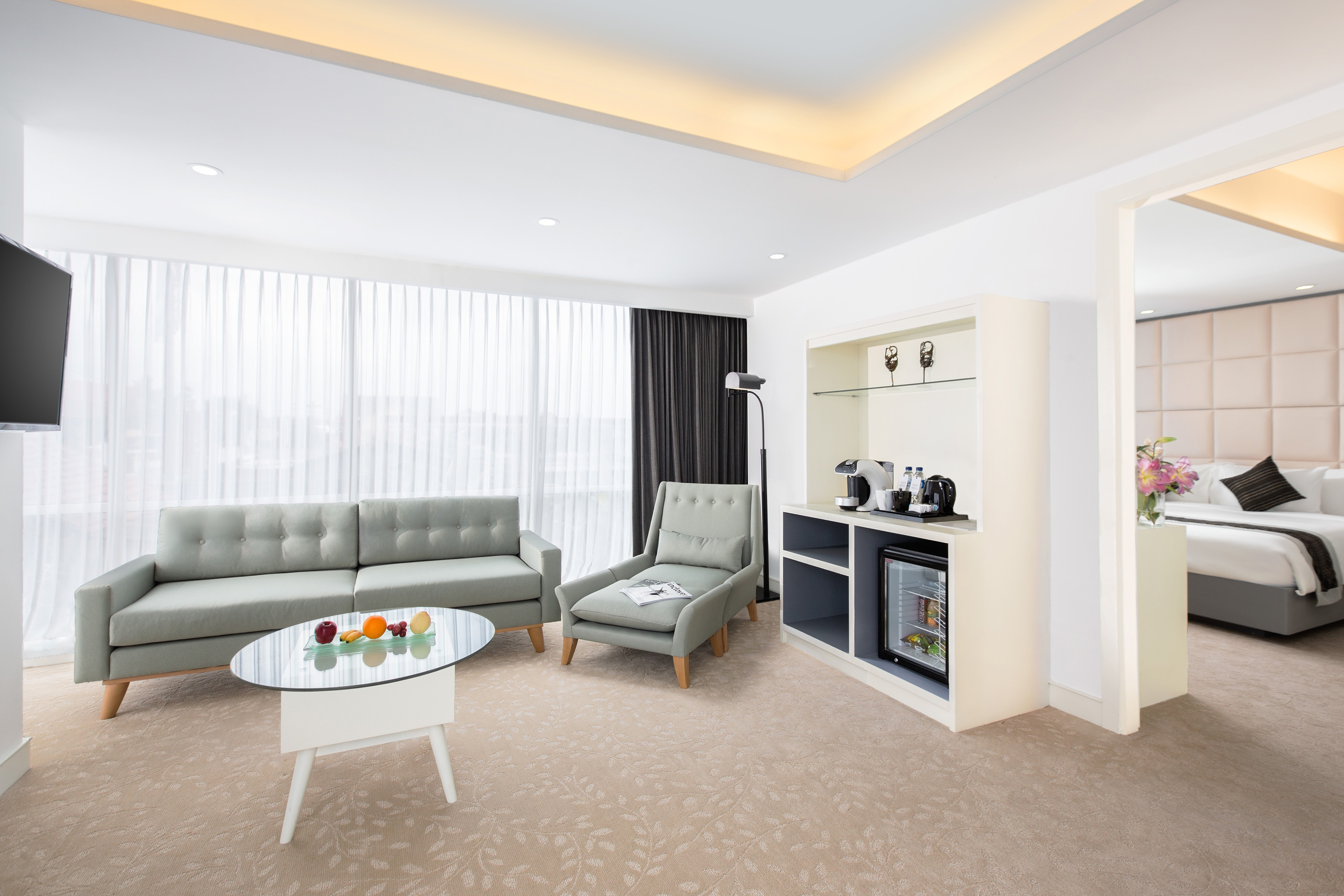 Contemporary hotel suite living space with sofa, armchair, mini-bar, and bedroom.
