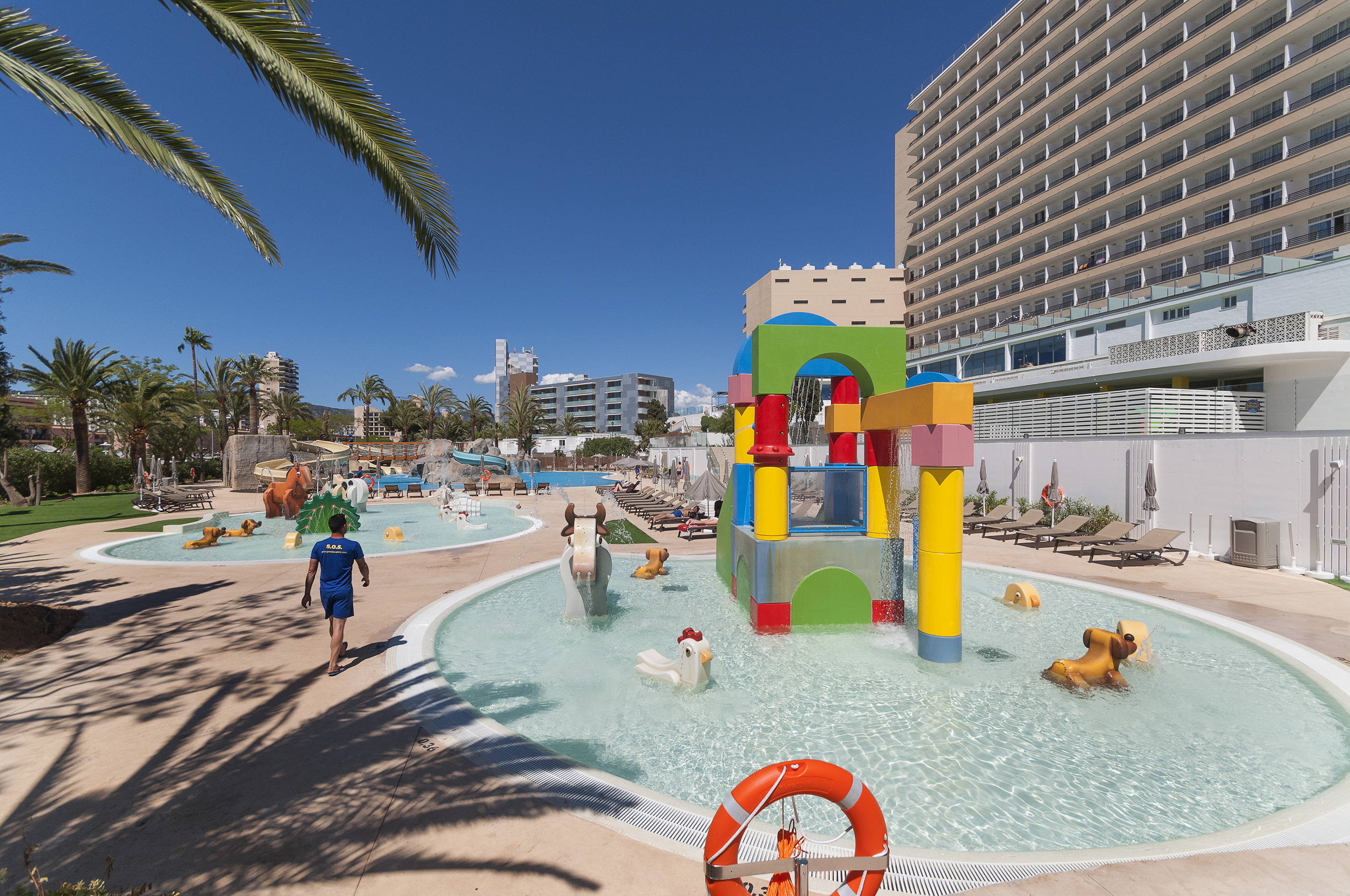 Sunny resort kids' pool with colorful water play structure, animal floats, hotel.