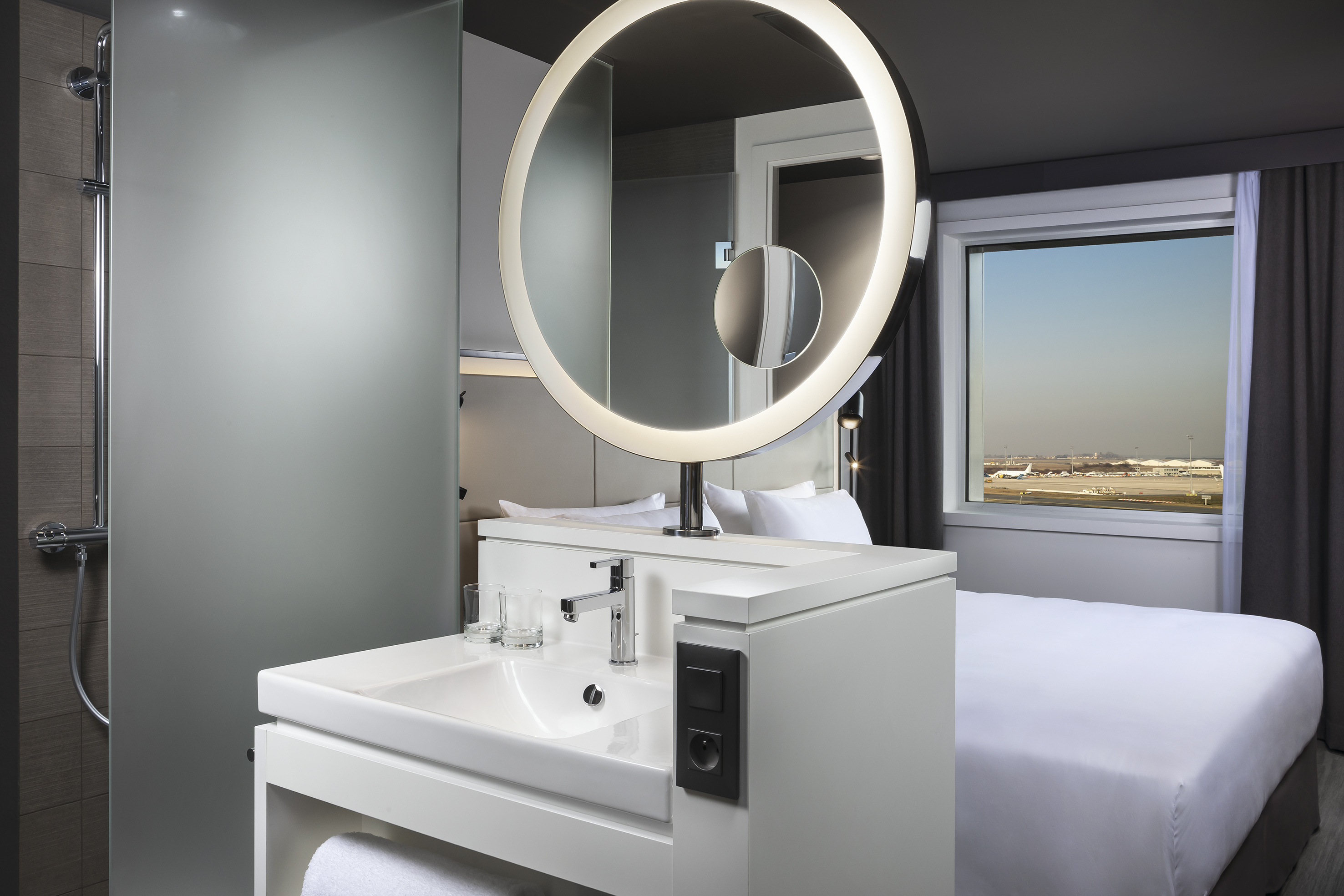 Modern hotel room with sink, illuminated mirror, bed, and airport view.