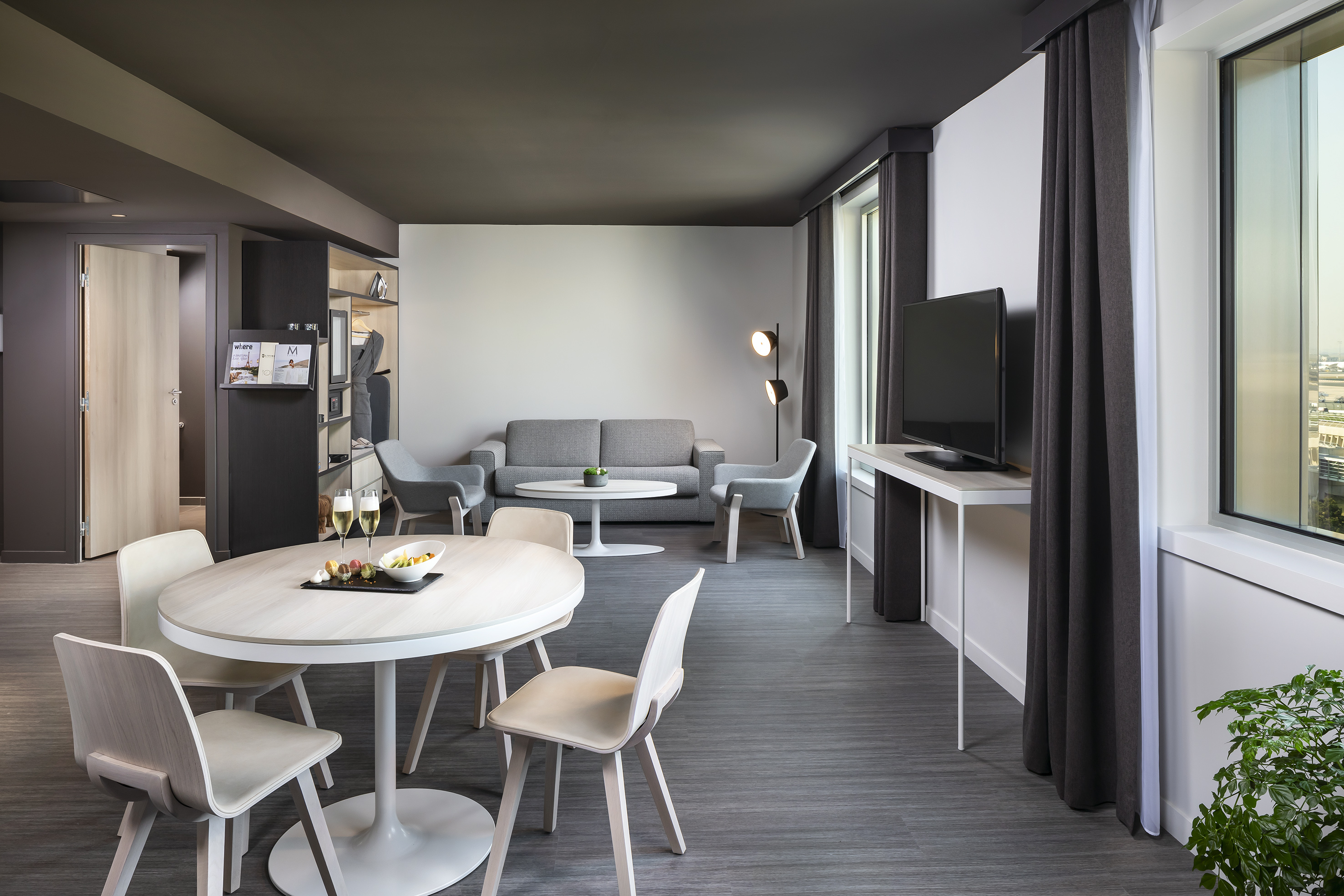 Modern hotel suite featuring dining area, grey sofa lounge, and large windows.