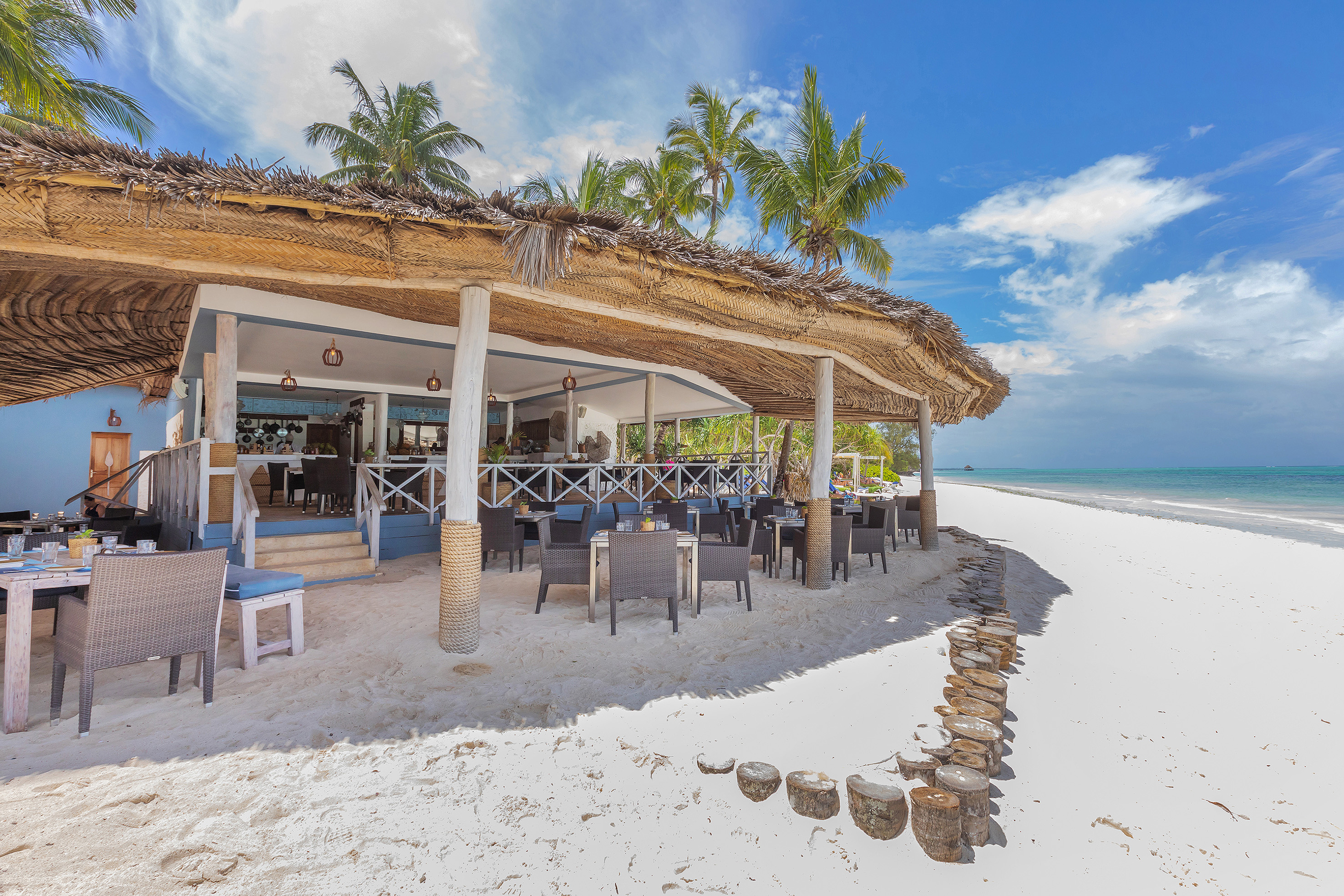 Beachfront restaurant with thatched roof, palm trees, white sand, and ocean.