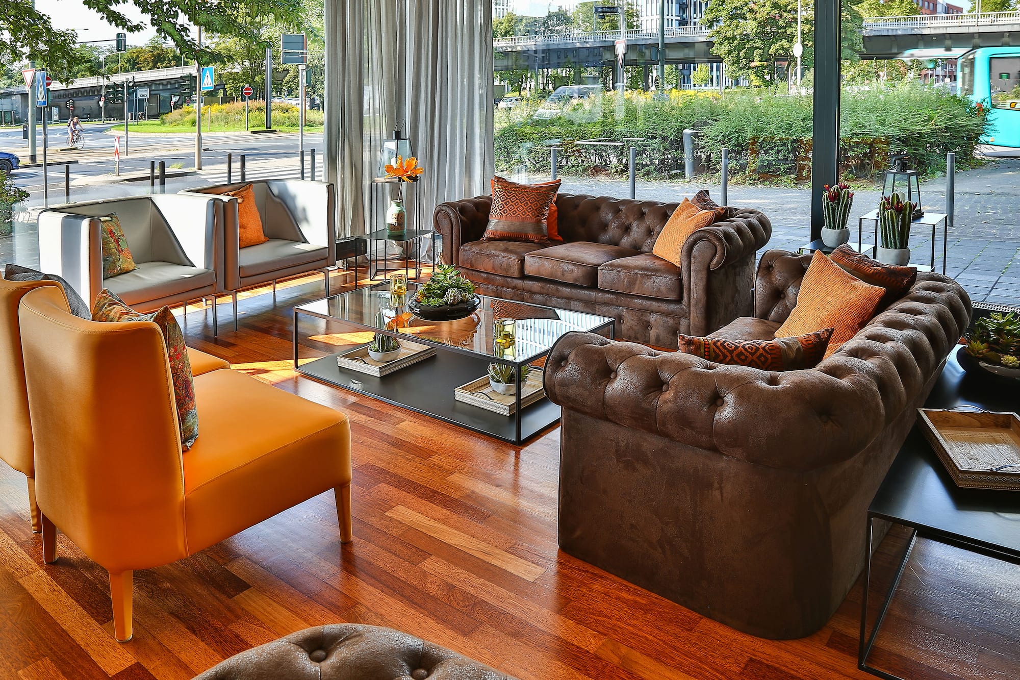 Modern lounge with brown tufted sofas, orange chairs, glass tables, and city view.
