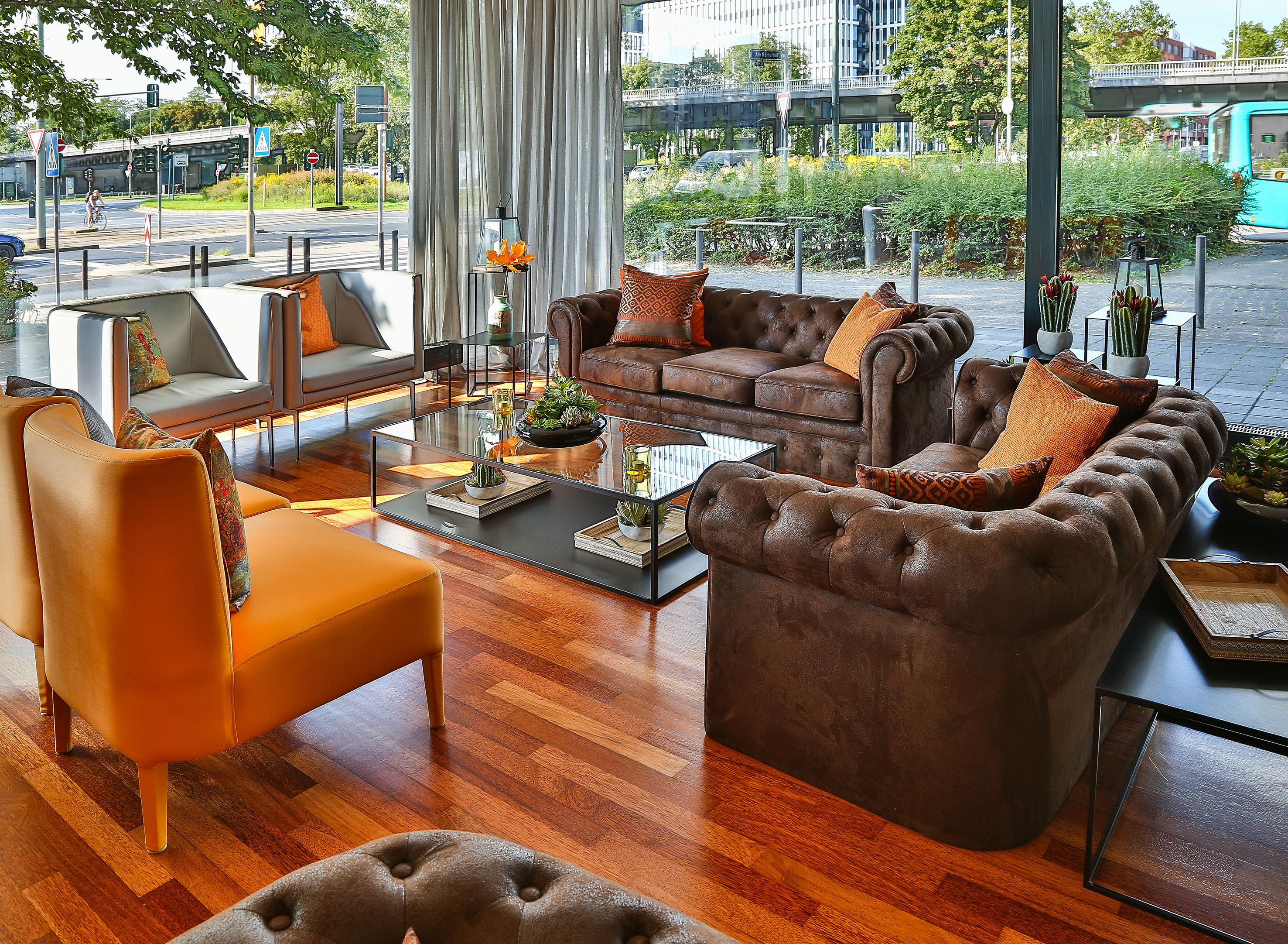 Modern lounge with brown tufted sofas, orange chairs, glass tables, and city view.