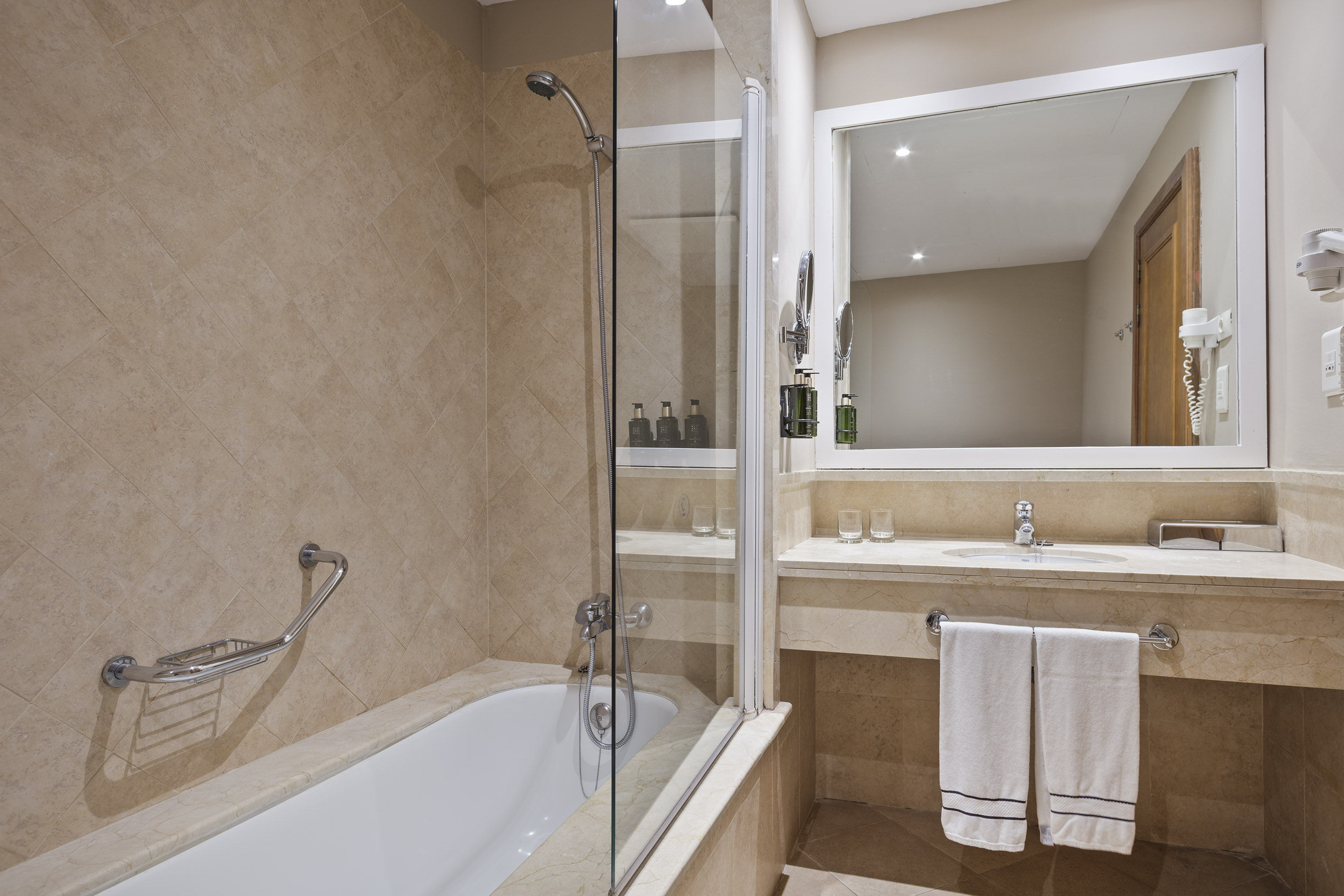 Modern hotel bathroom with beige tiles, bathtub, glass shower, vanity, and mirror.