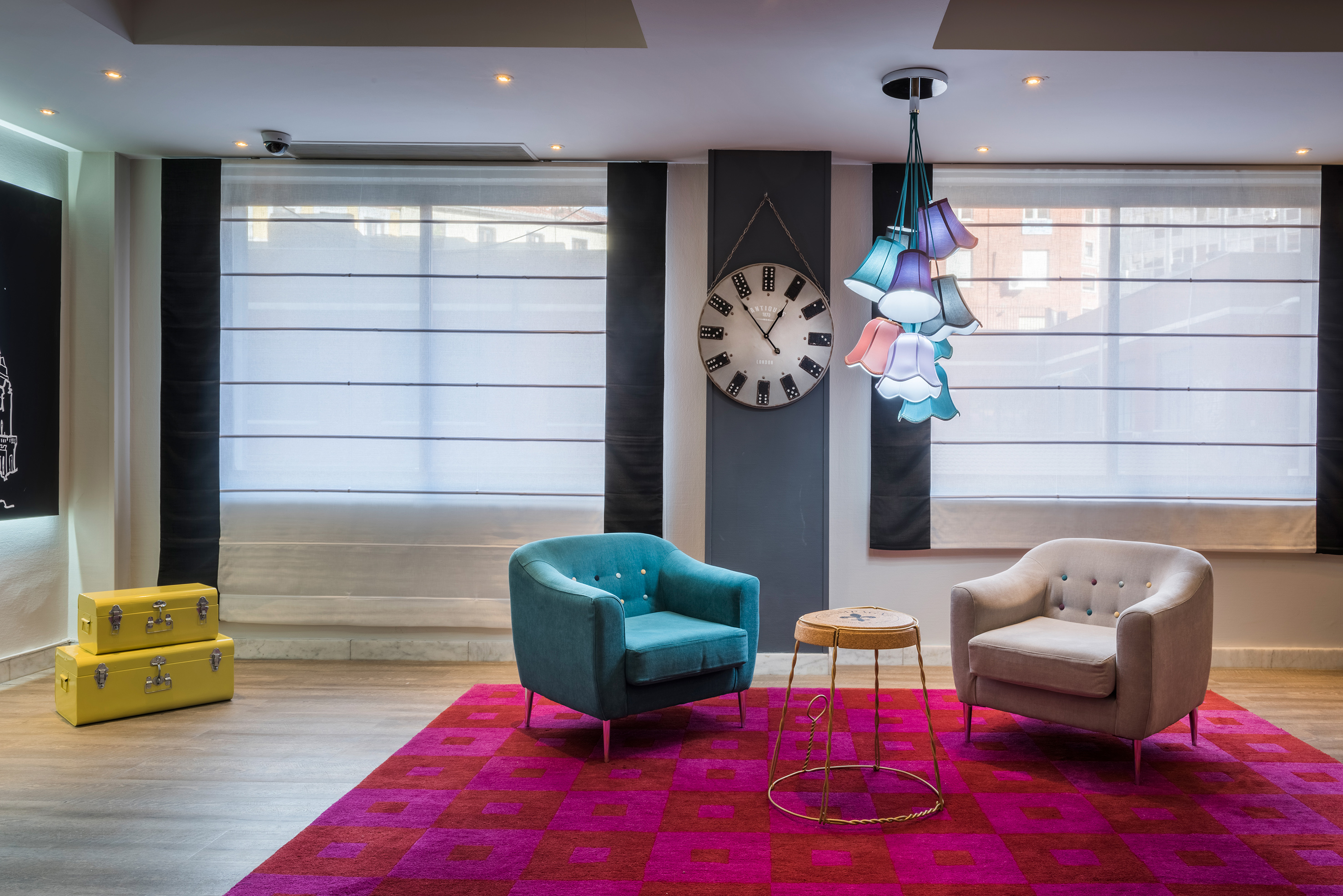 Vibrant modern lounge with teal and beige armchairs, colorful chandelier, red rug.