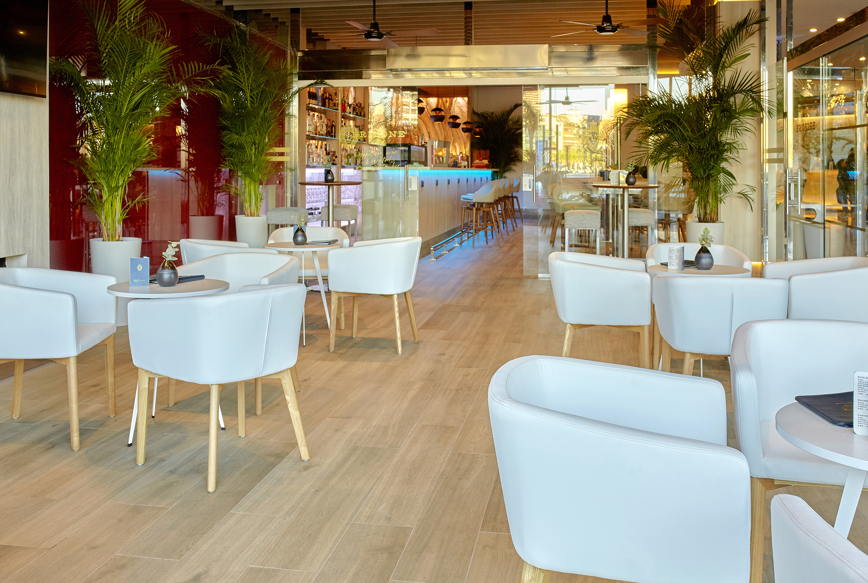 Modern hotel lounge bar with white chairs, palm plants, and illuminated bar counter.