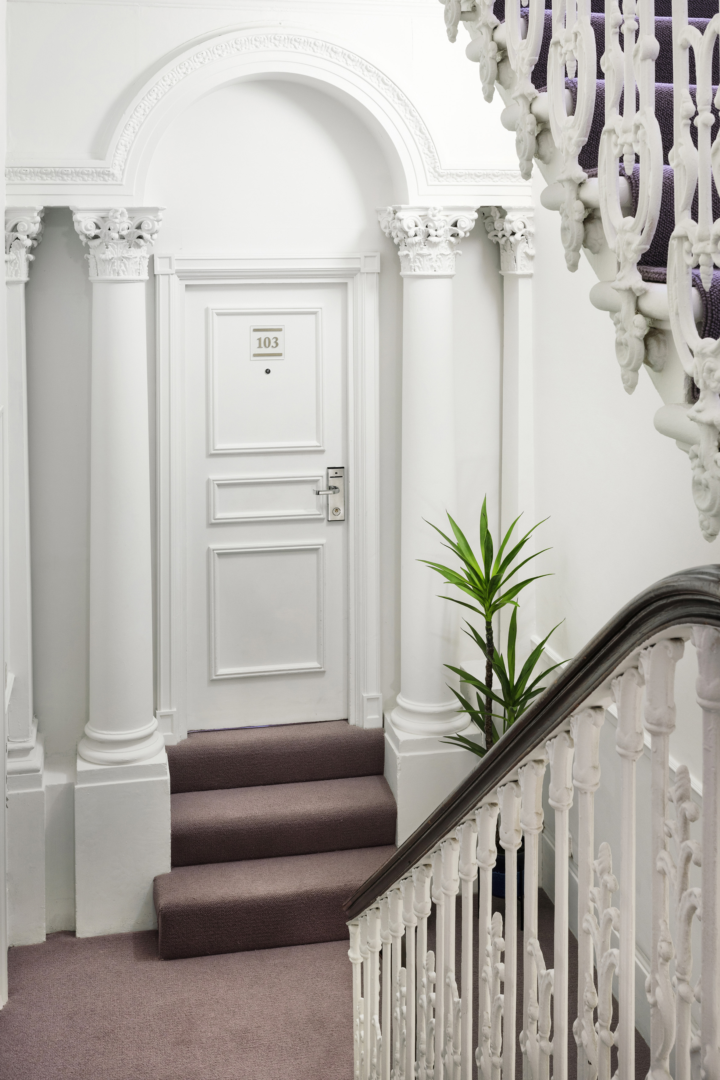 White classical hotel hallway: Room 103 door, columns, ornate staircase.
