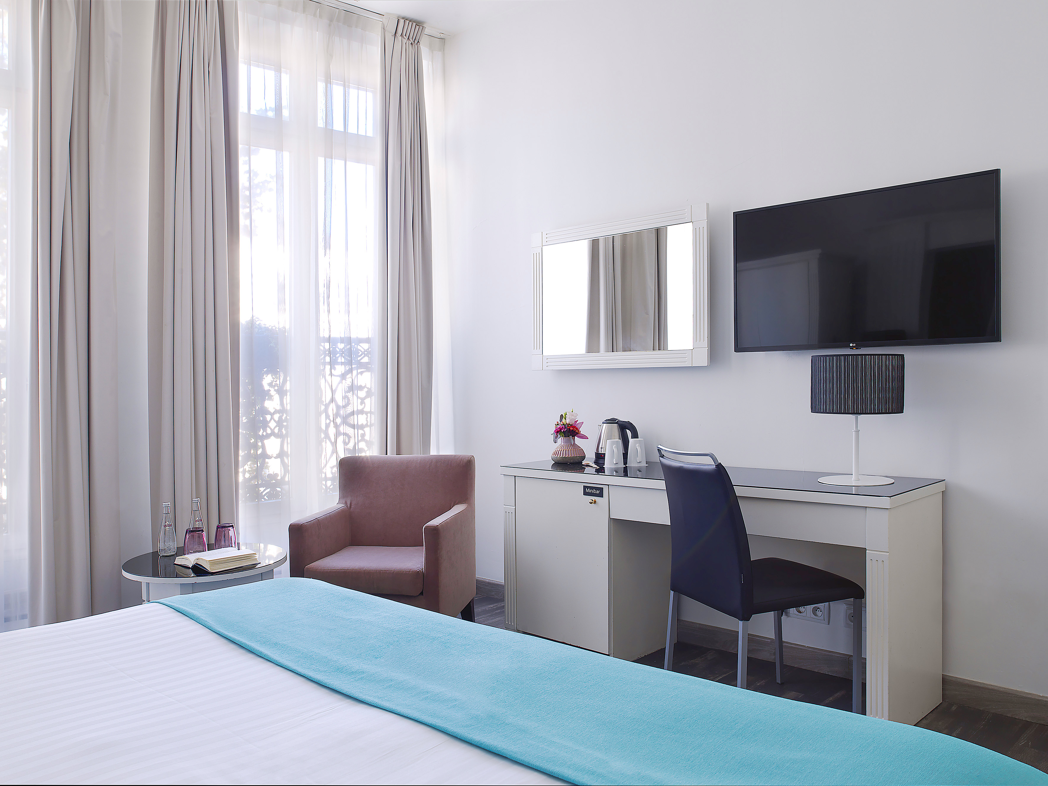 Modern hotel room with bed, teal runner, desk, TV, and bright window.