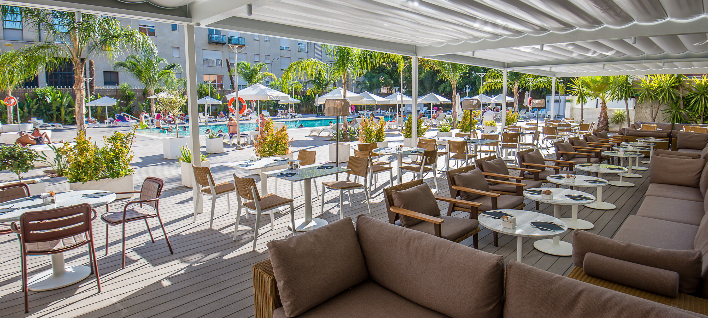 Outdoor resort pool terrace, shaded lounge seating, dining, palm trees.