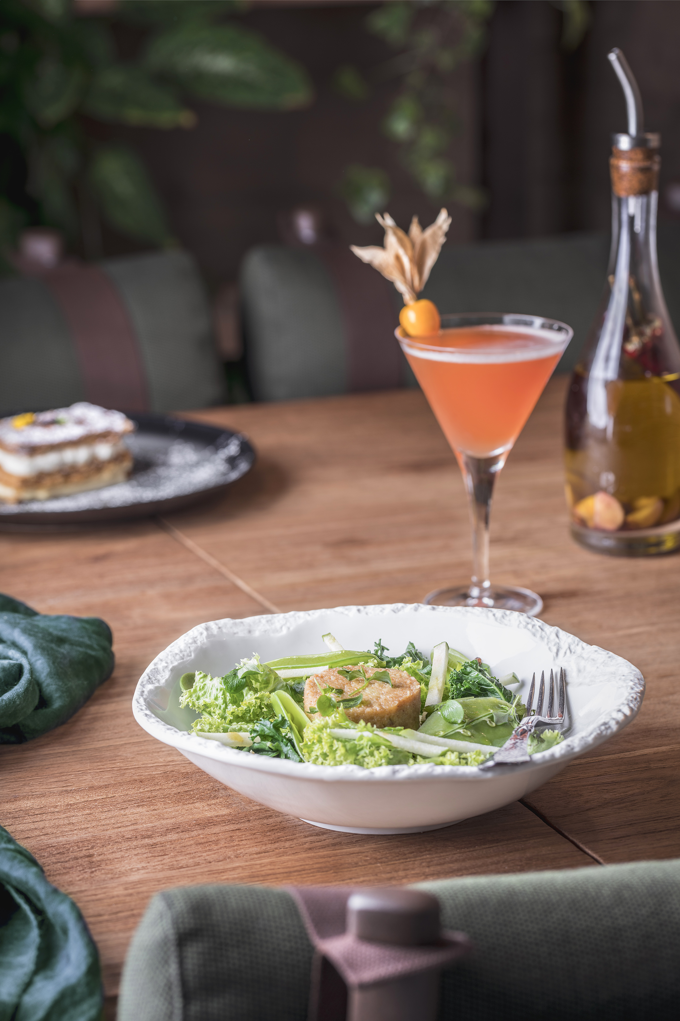 Green salad with fritter, pink cocktail, and dessert on a wooden restaurant table.