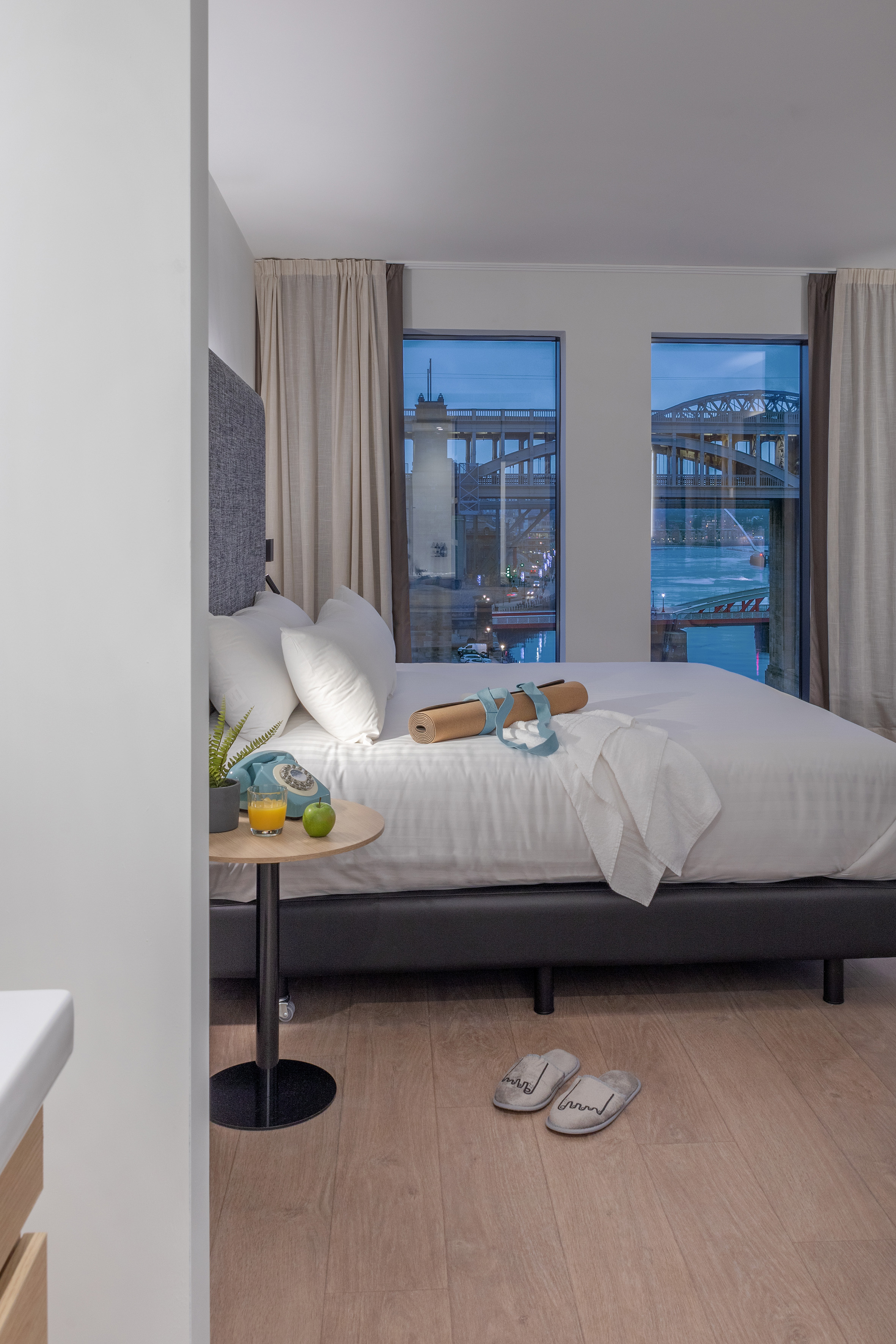 Modern hotel bedroom with city view, white bed, yoga mat, and nightstand.