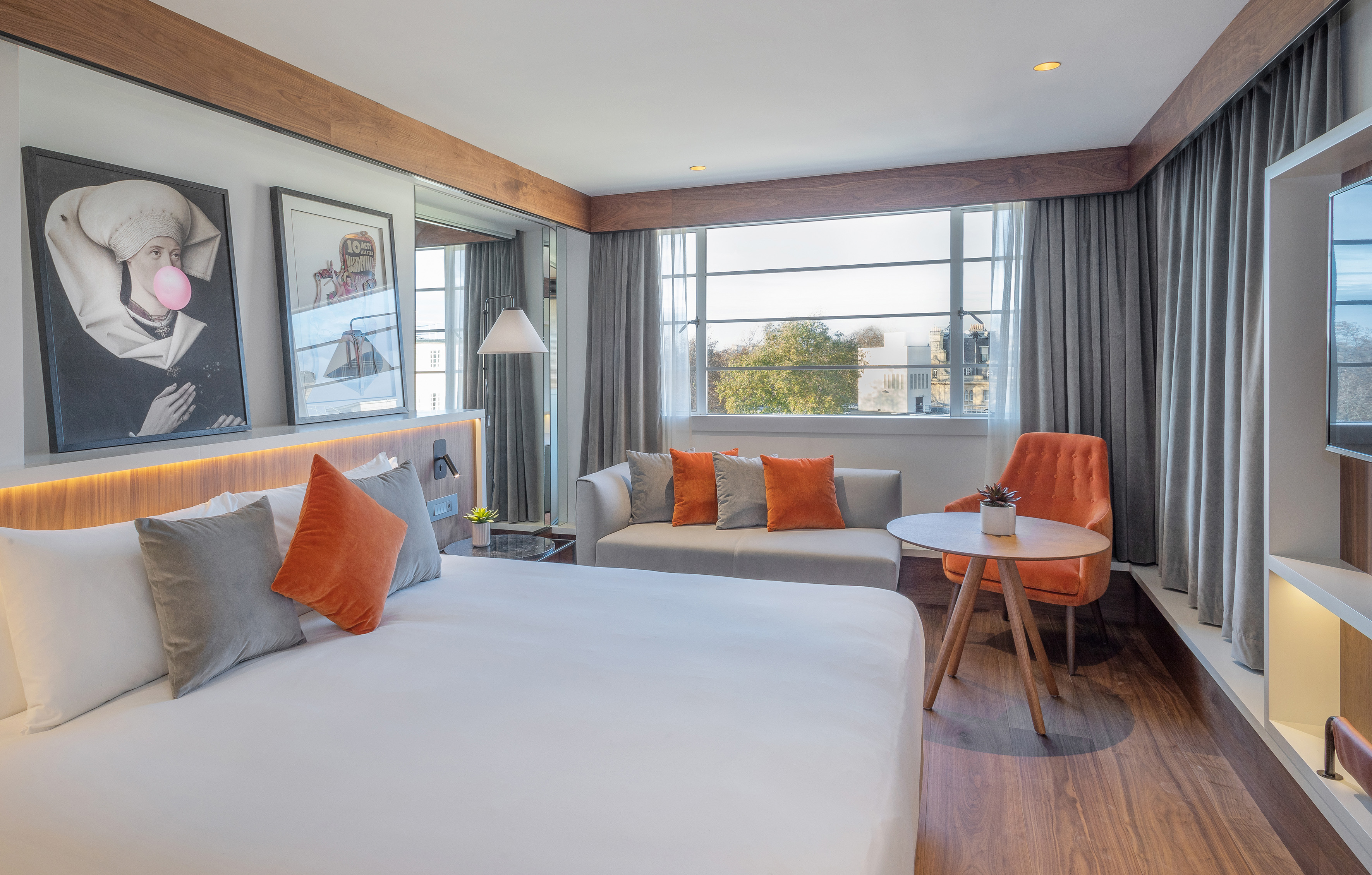 Modern hotel room with large bed, sofa, orange armchair, and window view.