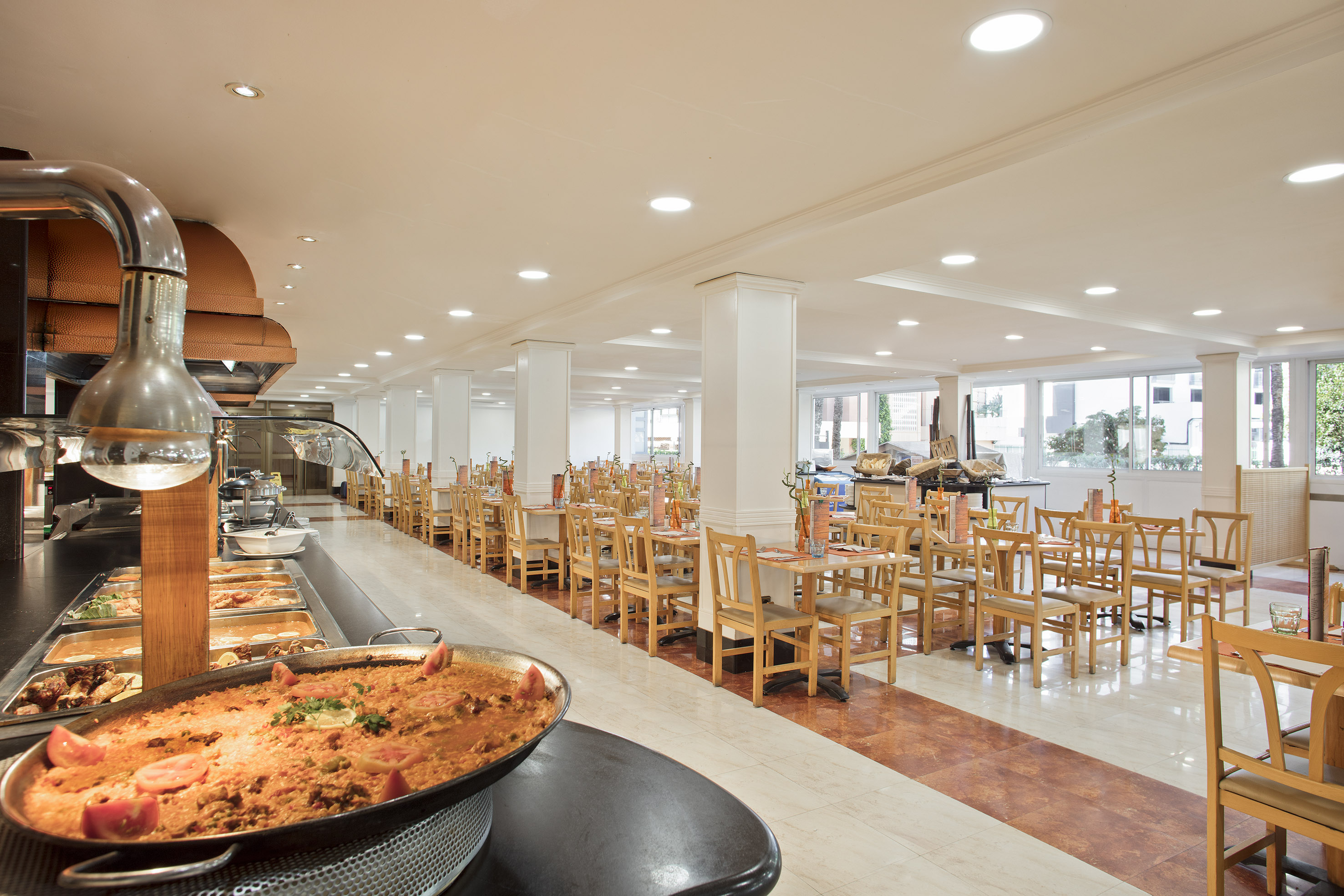 Bright, spacious hotel buffet with paella and empty dining tables.
