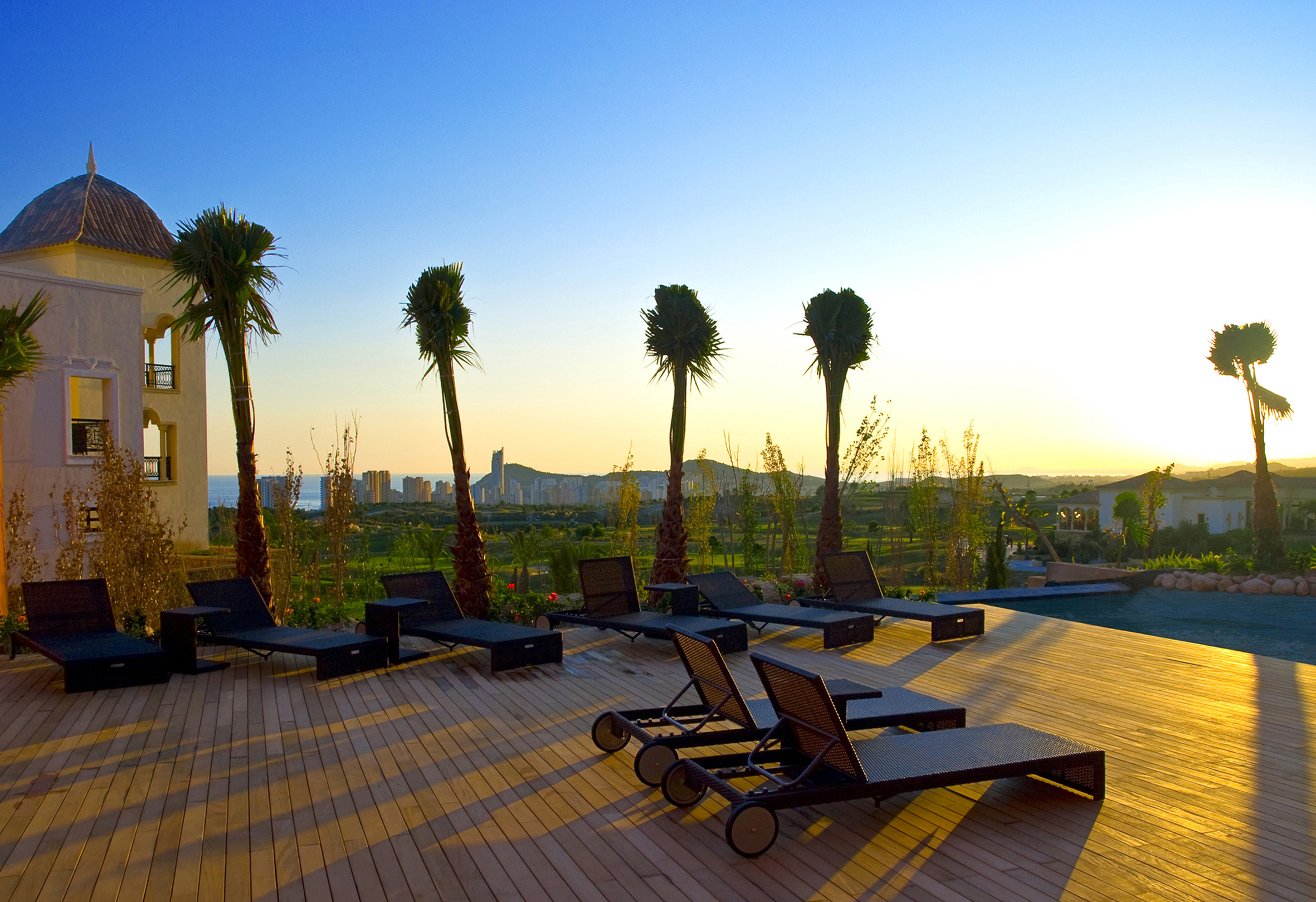 Sunset over resort deck with sun loungers, palm trees, and city skyline.